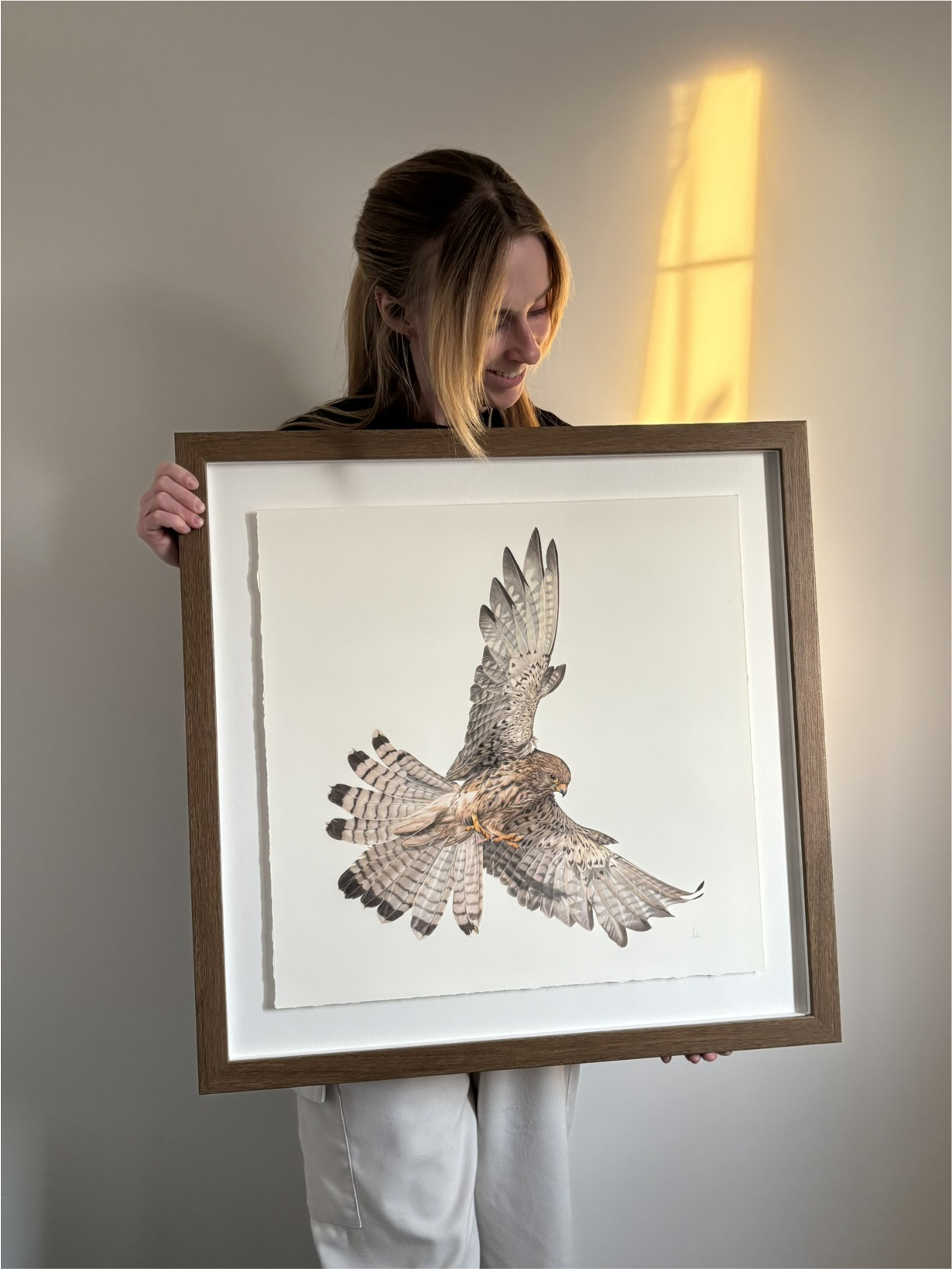 Woman holding a framed illustration of a bird with distinctive feather patterns, smiling as she looks down at it.