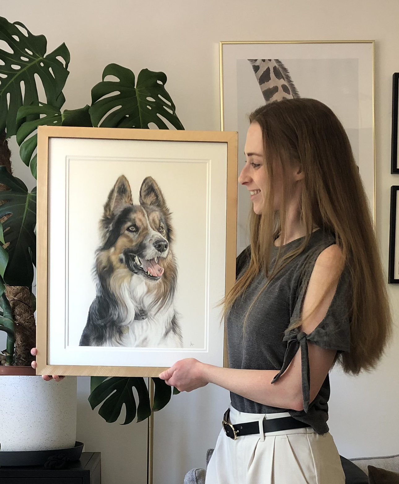 A woman with long red hair holding a framed portrait of a Border Collie dog, smiling while looking at the artwork.