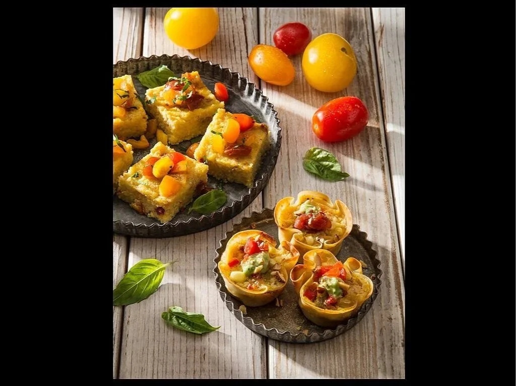 Various small savory dishes on round metal plates, including polenta squares topped with vegetables and mini pasta cups filled with vegetables, on a wooden surface with fresh cherry and yellow tomatoes and basil leaves scattered around.