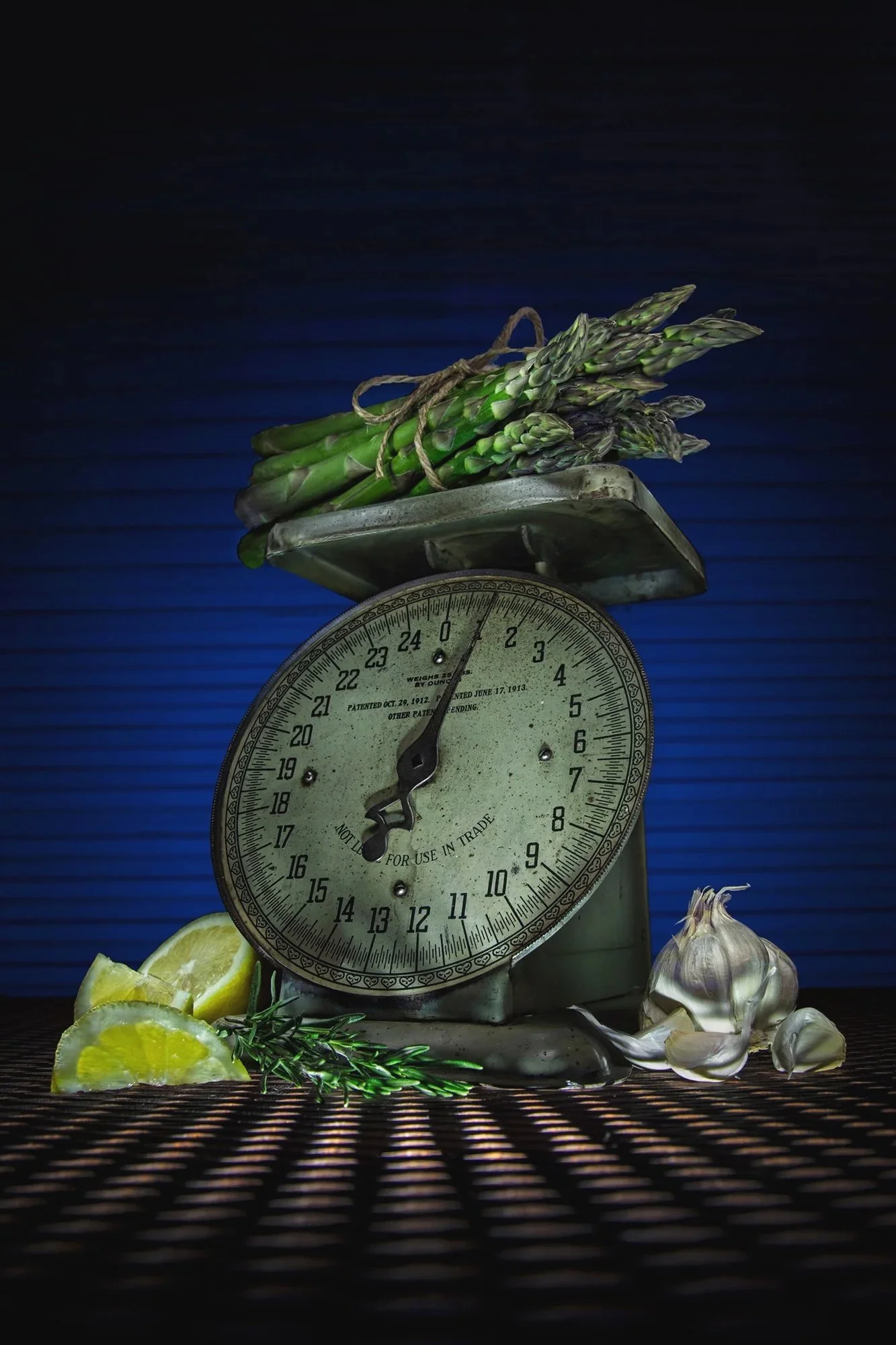 A vintage kitchen scale with a bunch of fresh asparagus on top, lemon wedges and garlic cloves in front, and a sprig of rosemary beside.