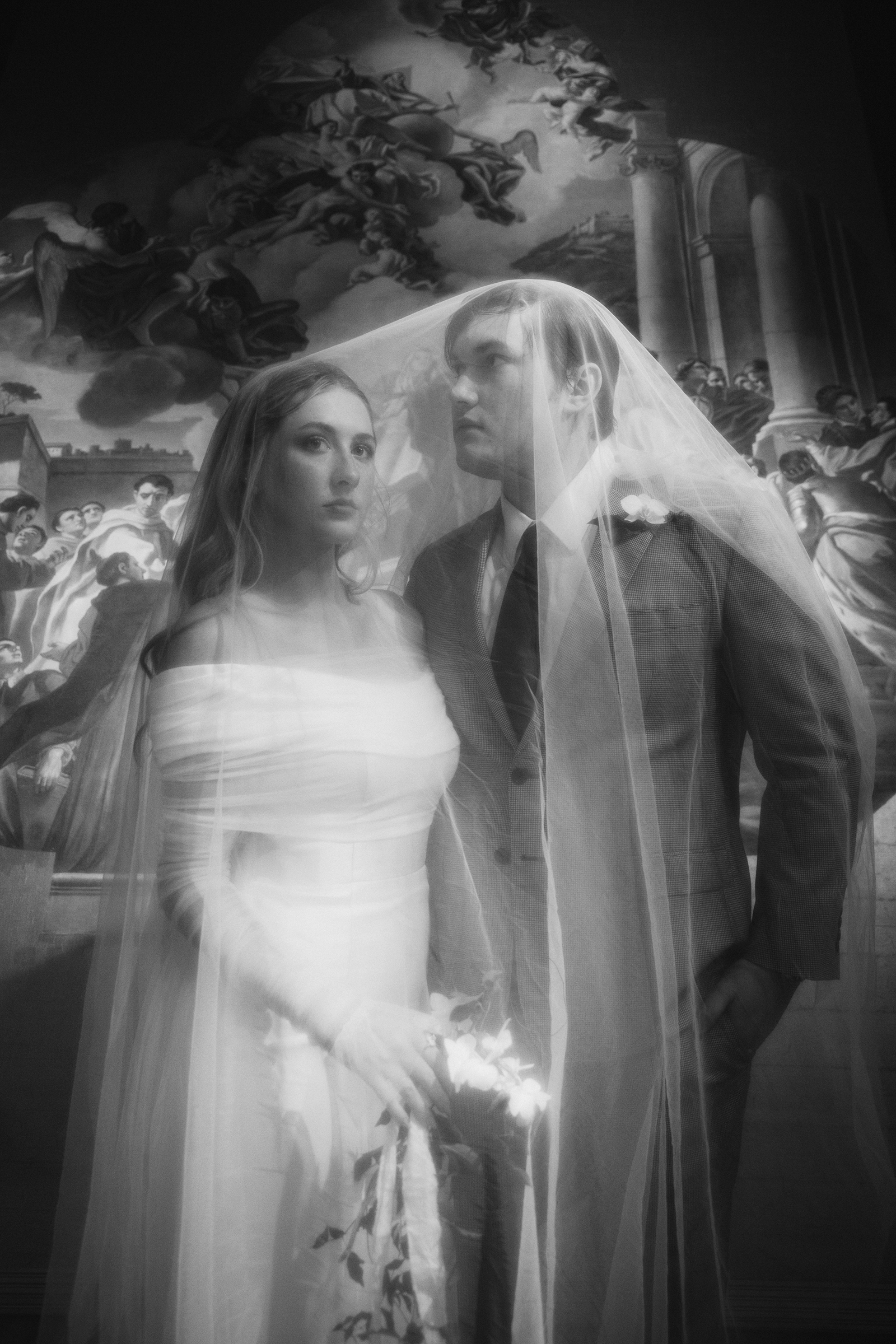 Black and white photo of a bride and groom standing close, with a painted background depicting a classical scene with angels and people. The bride is wearing a strapless wedding dress and holding flowers, while the groom is in a suit and tie, with a veil over both.