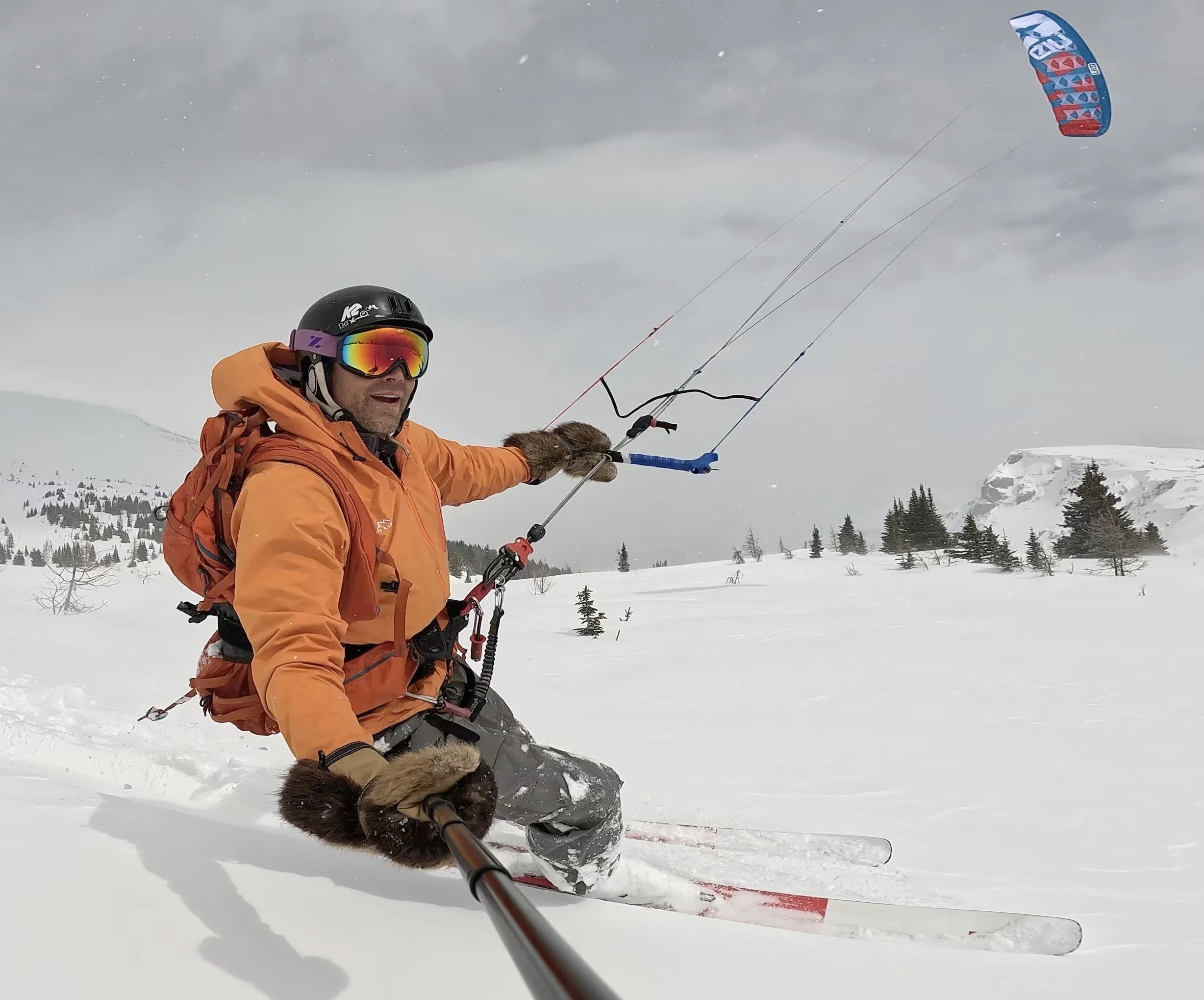 A person snowboarding in a snowy mountain landscape wearing an orange jacket, black helmet, reflective goggles, and holding a selfie stick while taking a photo. Sunshine Meadows. Gin Kiteboarding. Shaman 3.1