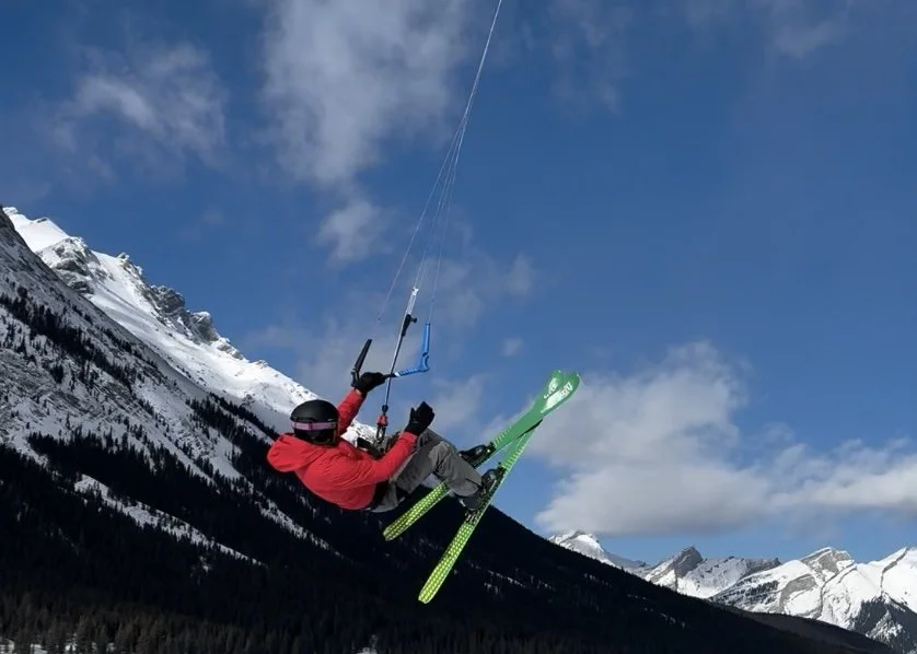Person in red jacket and gray pants riding a green ski kite in a snowy mountain landscape. Snow kiting Spray Lakes, Alberta. Canada. 