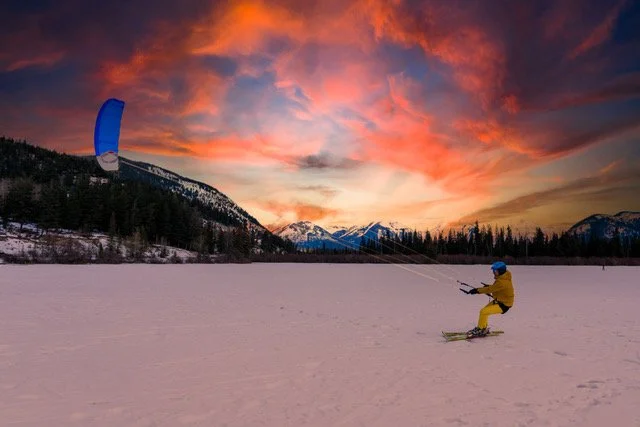 Person in yellow winter gear snowkiting on a frozen lake at sunset with colorful sky and mountains in the background.