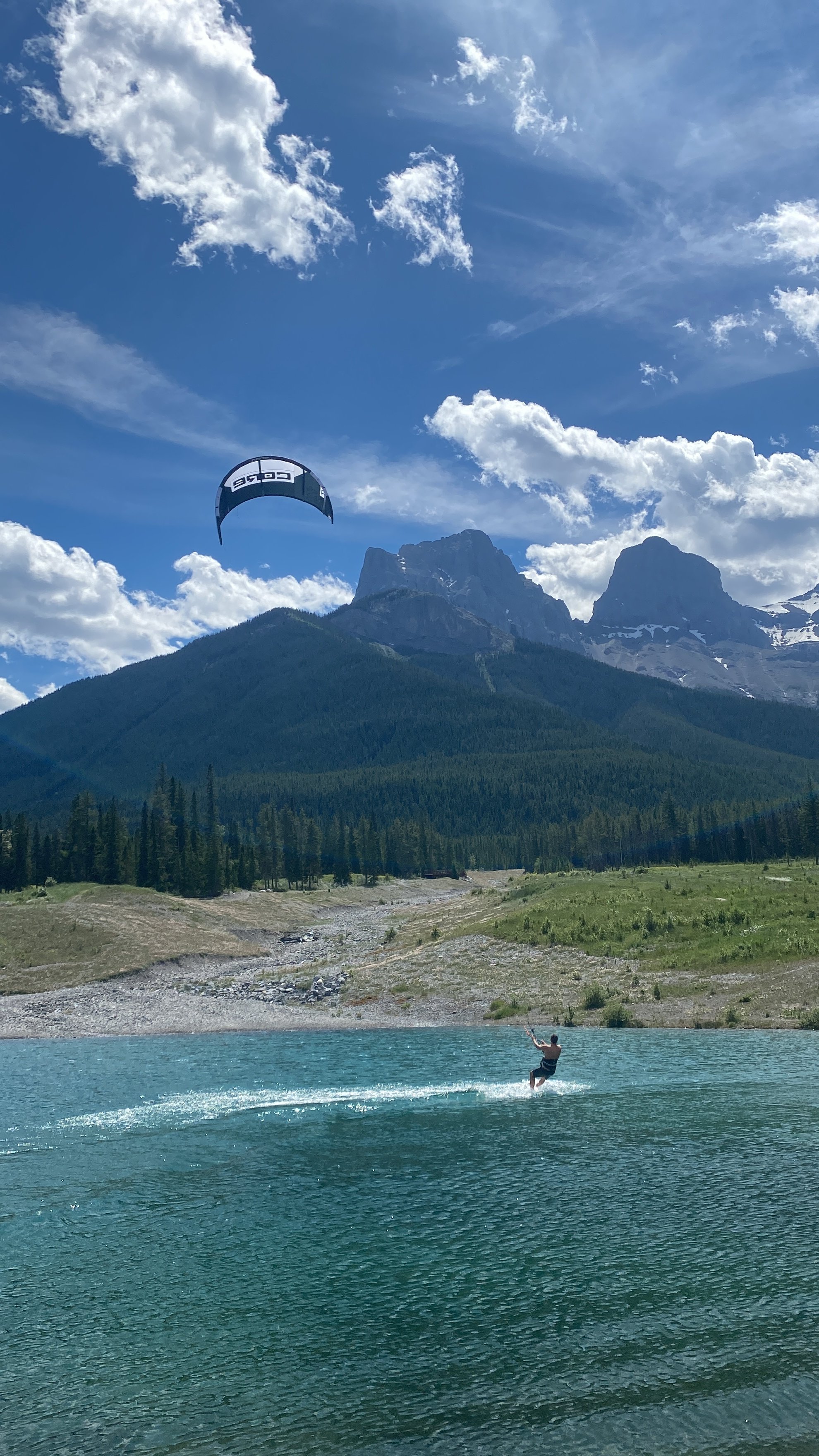 A person waterskiing on a large body of turquoise water with mountains and trees in the background, and a kite in the sky.