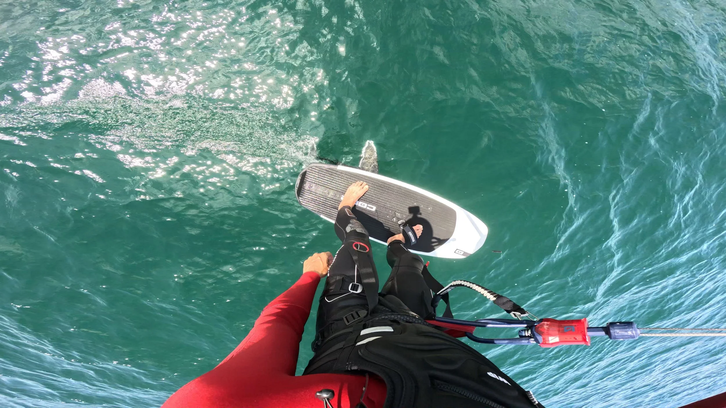 Person standing on a Foil board on green water, wearing black wetsuit and red jacket, holding the paddle, with a small propeller attached at the back. Core SLC Foil Package. Core Kiteboarding.