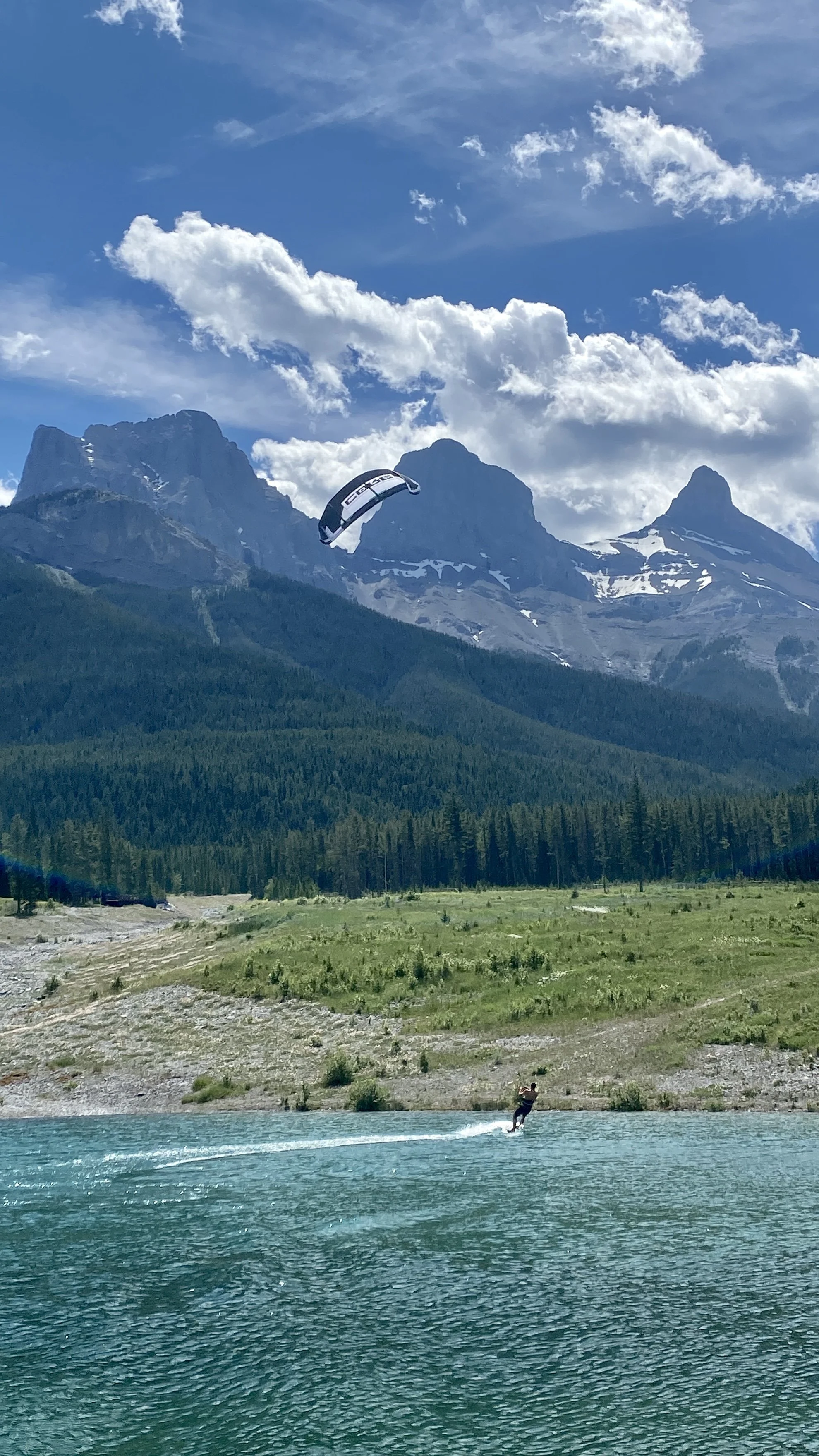 A person kite surfing on a turquoise lake with mountains, green forests, and a partly cloudy sky in the background.