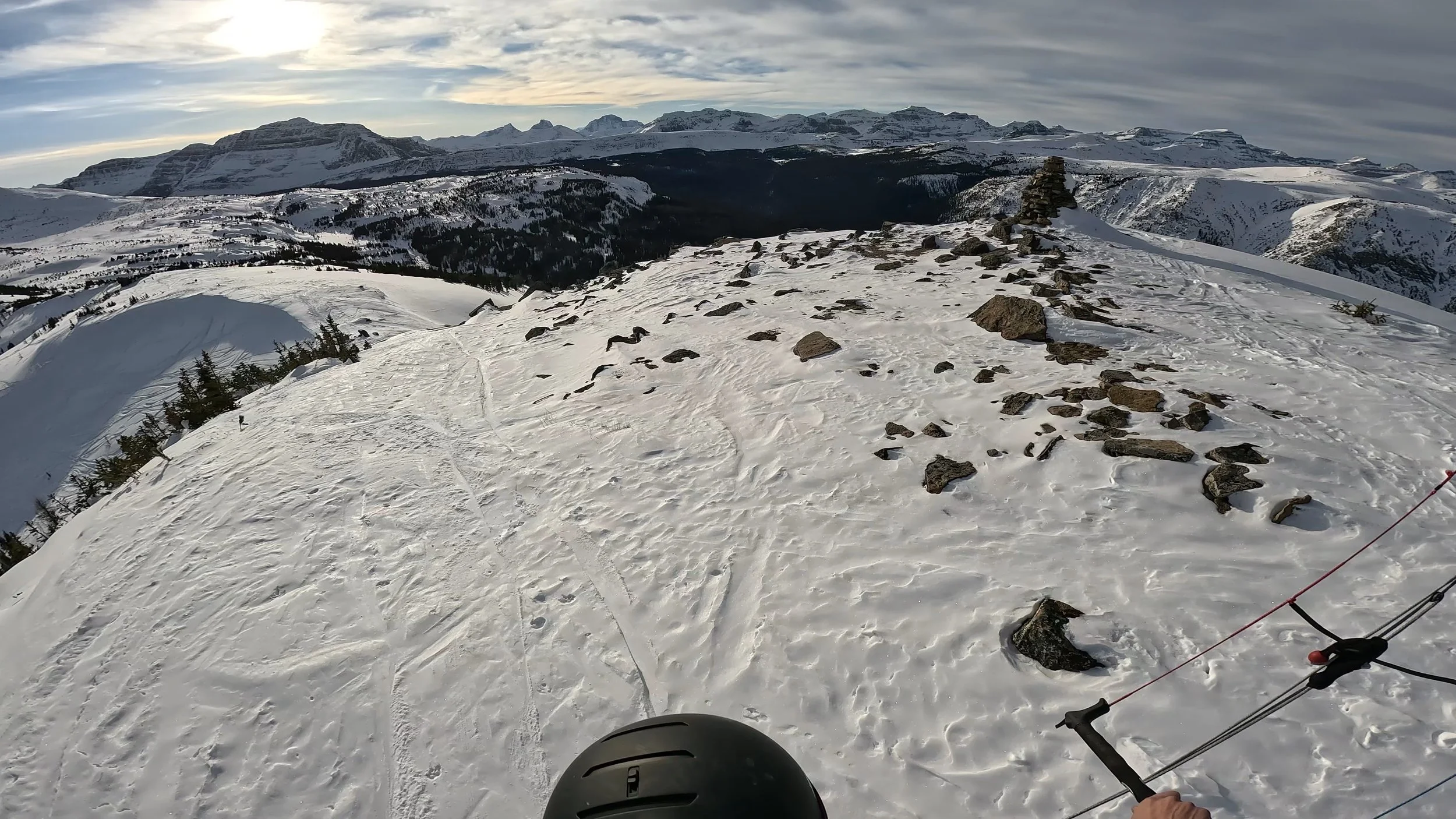 Snow-covered mountain landscape with rocky terrain and a trail, view from a skier or snowboarder with a helmet and ski poles, under a cloudy sky.