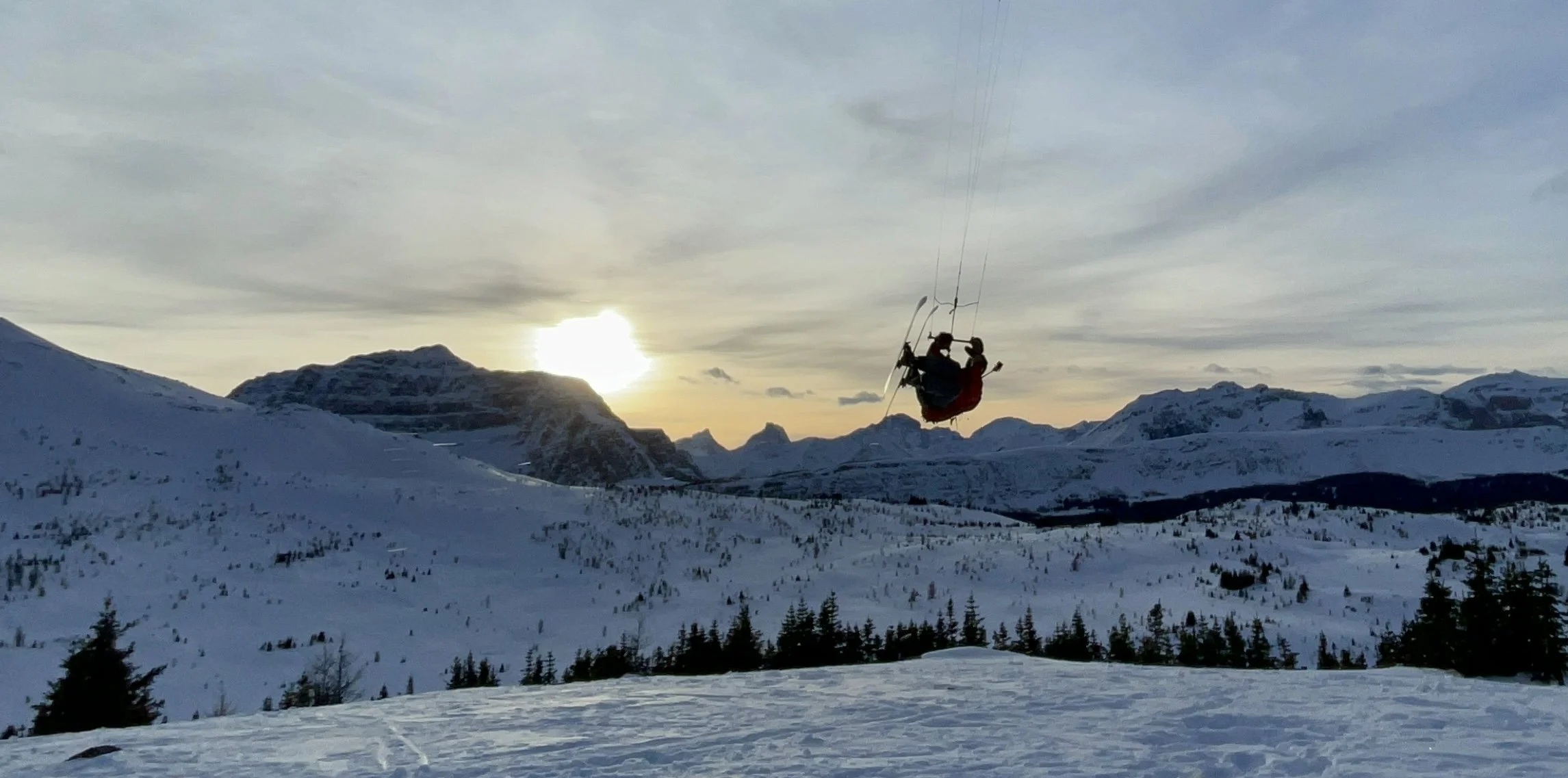 A person paragliding over snow-covered mountains and trees at sunset.