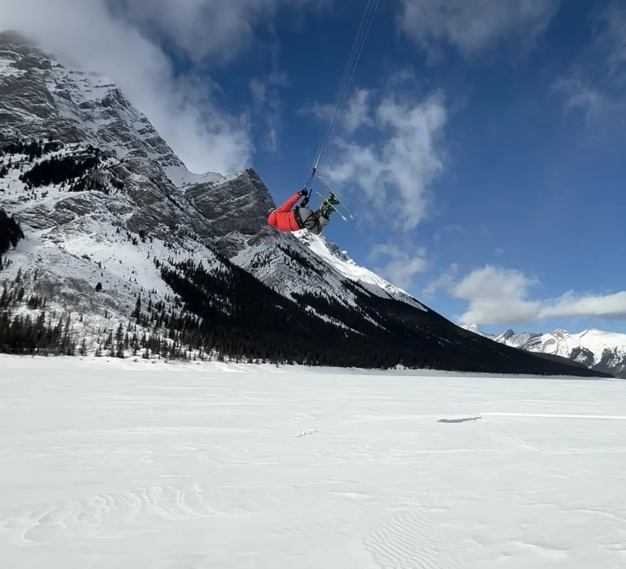 Person in red jacket kite skiing across a snowy mountain landscape with snow-covered mountains and a partly cloudy sky in the background.