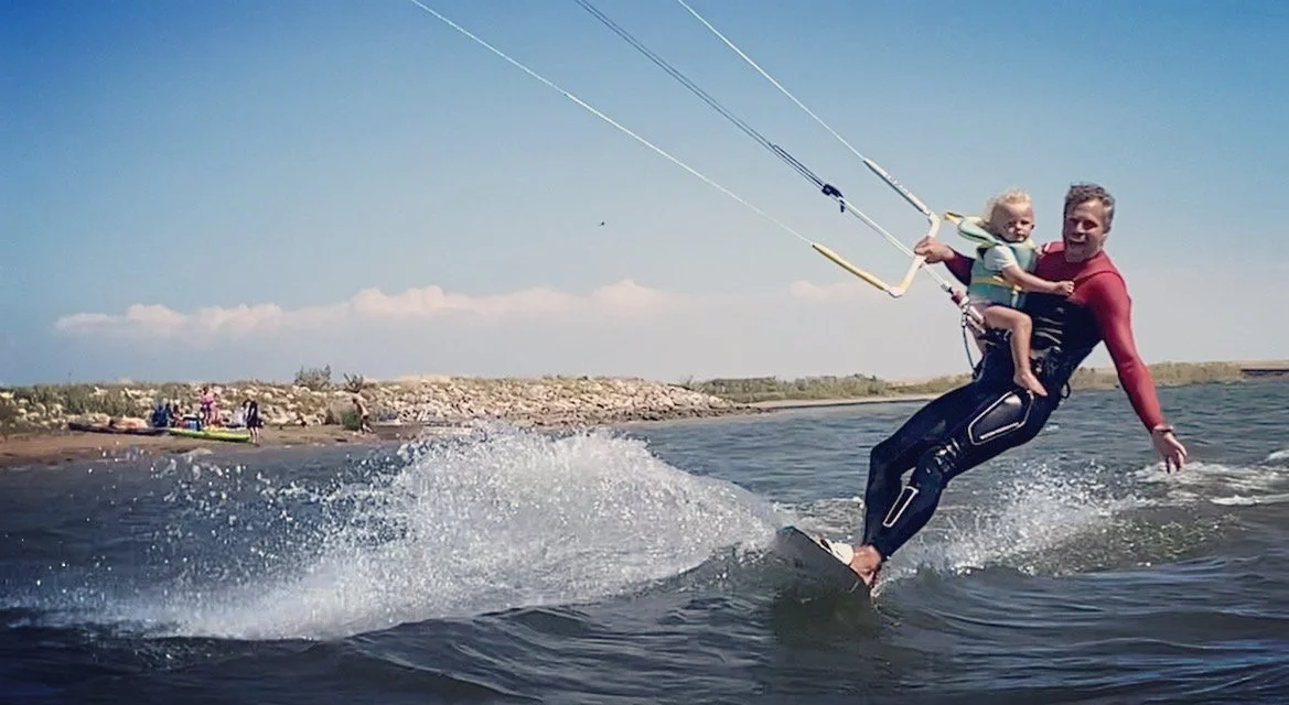 A man is kite surfing in the water with a young child sitting in a harness in front of him. The man is holding the kite's control bar, and they are gliding across the water, creating a splash. The shoreline with rocks and a few boats is visible in th