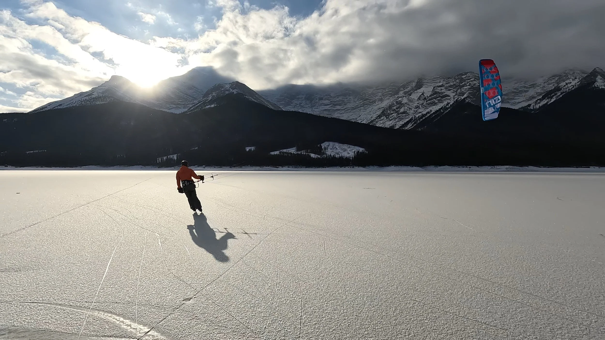 A person ice skating on a snow-covered frozen lake with snow-capped mountains in the background, under a partly cloudy sky with the sun shining.