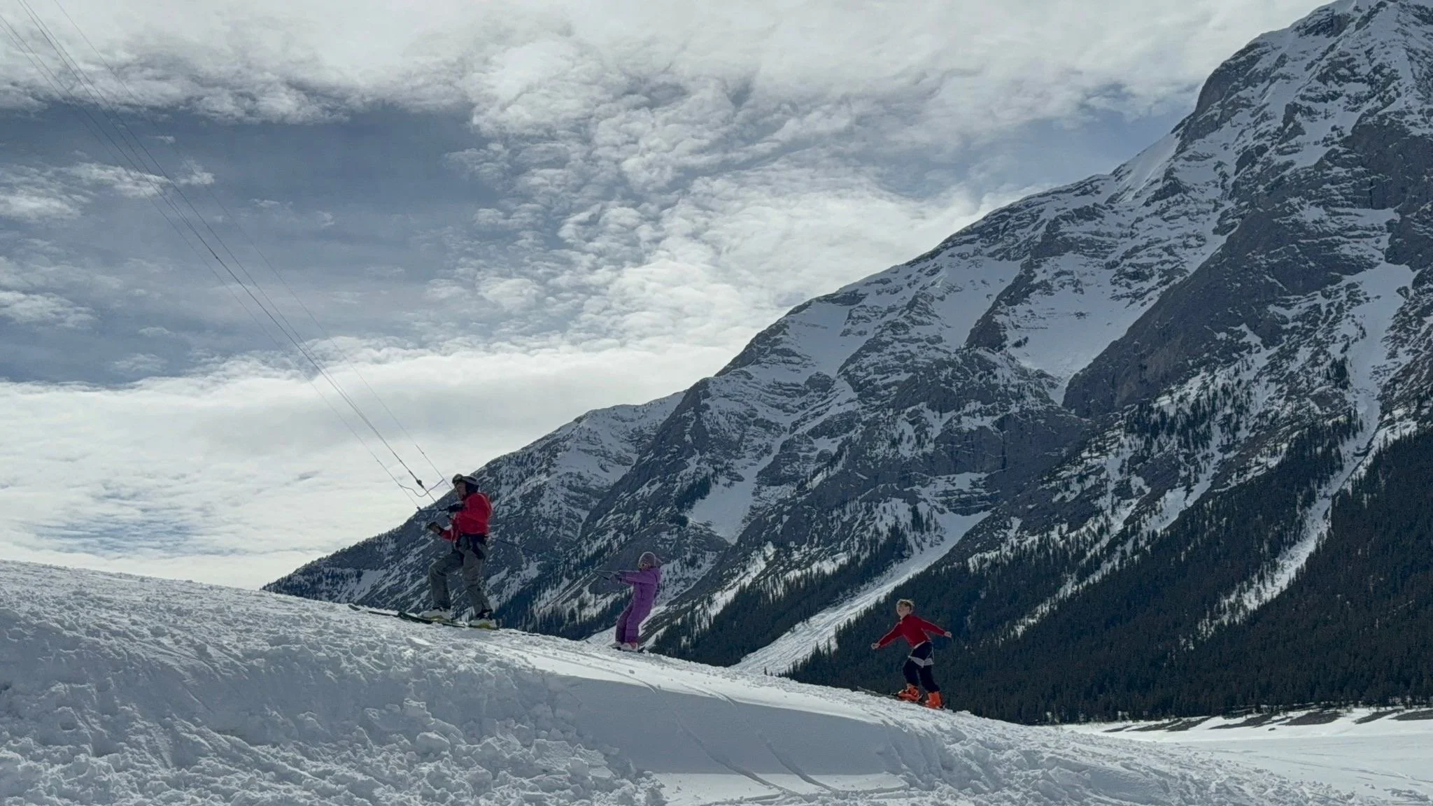 People skiing down a snowy mountain slope with tall snow-covered mountains and cloudy sky in the background.