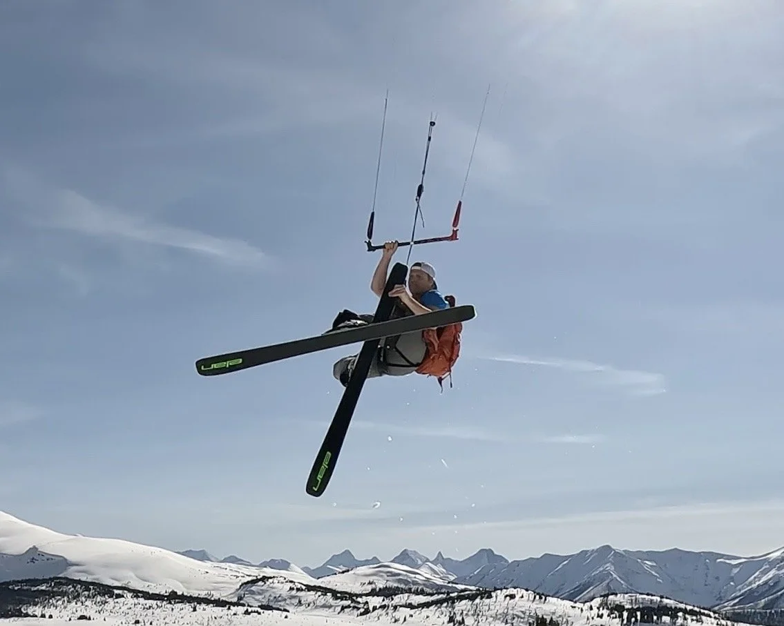 Person kitesurfing in the snow, lifted into the air with snow-covered mountains in the background. Snowkiting in Sunshine Meadows, Alberta.