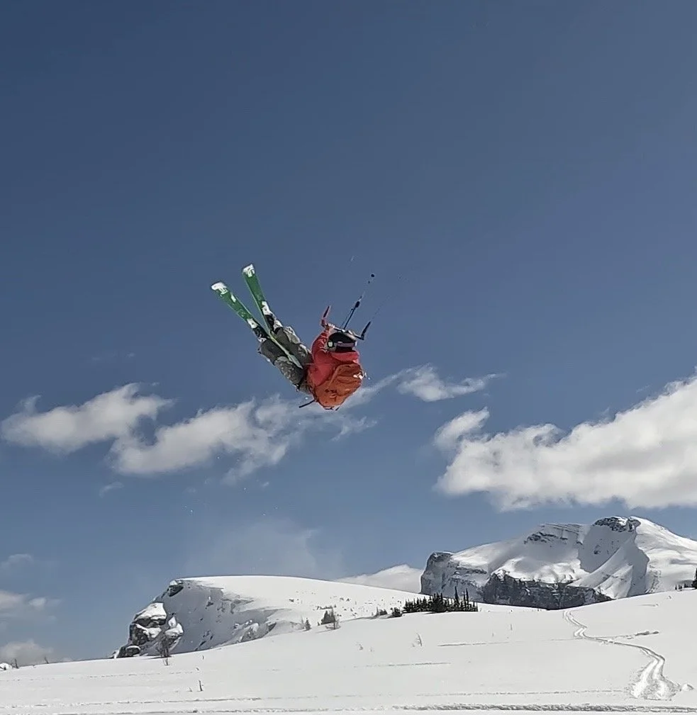 A skier performing a mid-air trick over snowy mountain terrain under a clear blue sky. Jumps at Sunshine Ski Resort. 