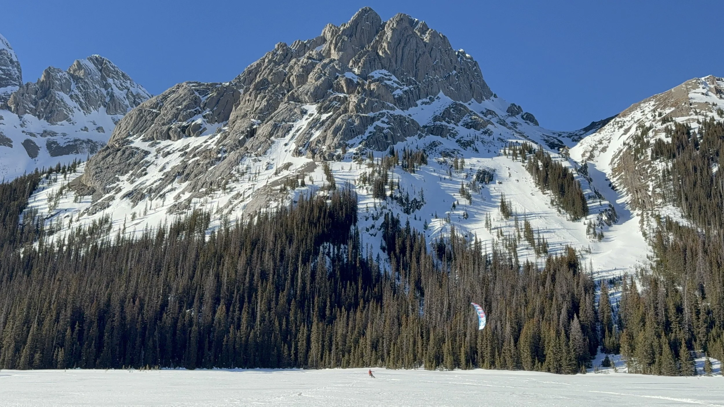Snow-covered mountain range with rugged peaks and a forested slope, clear blue sky, frozen lake or snowfield in the foreground with a person and a kite on the snow.