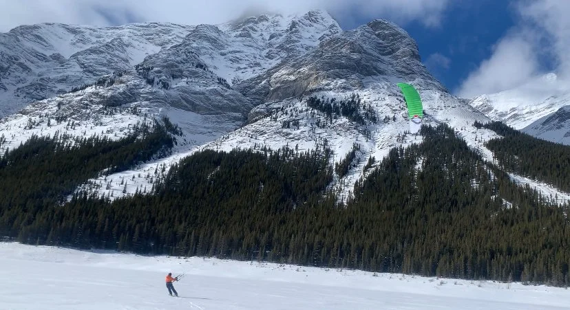 A person is cross-country skiing on snow with snowy mountains and a pine forest in the background. A green kite is flying in the sky above the mountains.