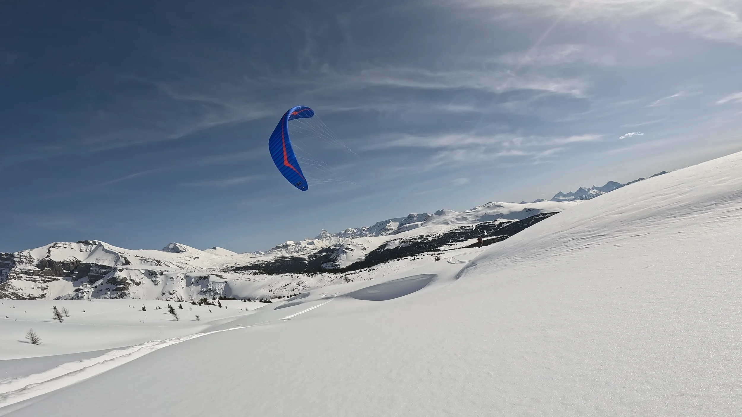 A snow-covered mountain landscape with a blue and red kite soaring in the sky. Tracks in the snow suggest recent activity, and distant mountains are visible on the horizon. Sunshine Meadows. The Meadows. Our favorite run. 