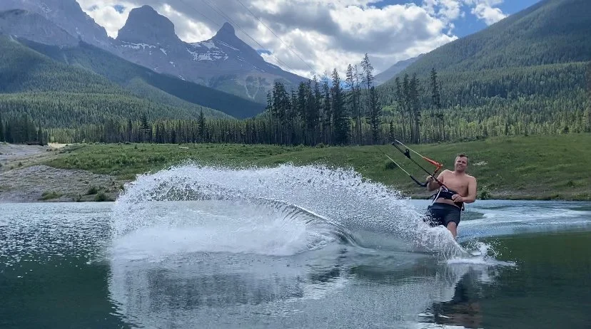 A person water skiing on a lake with mountains in the background, surrounded by forest and cloudy sky.