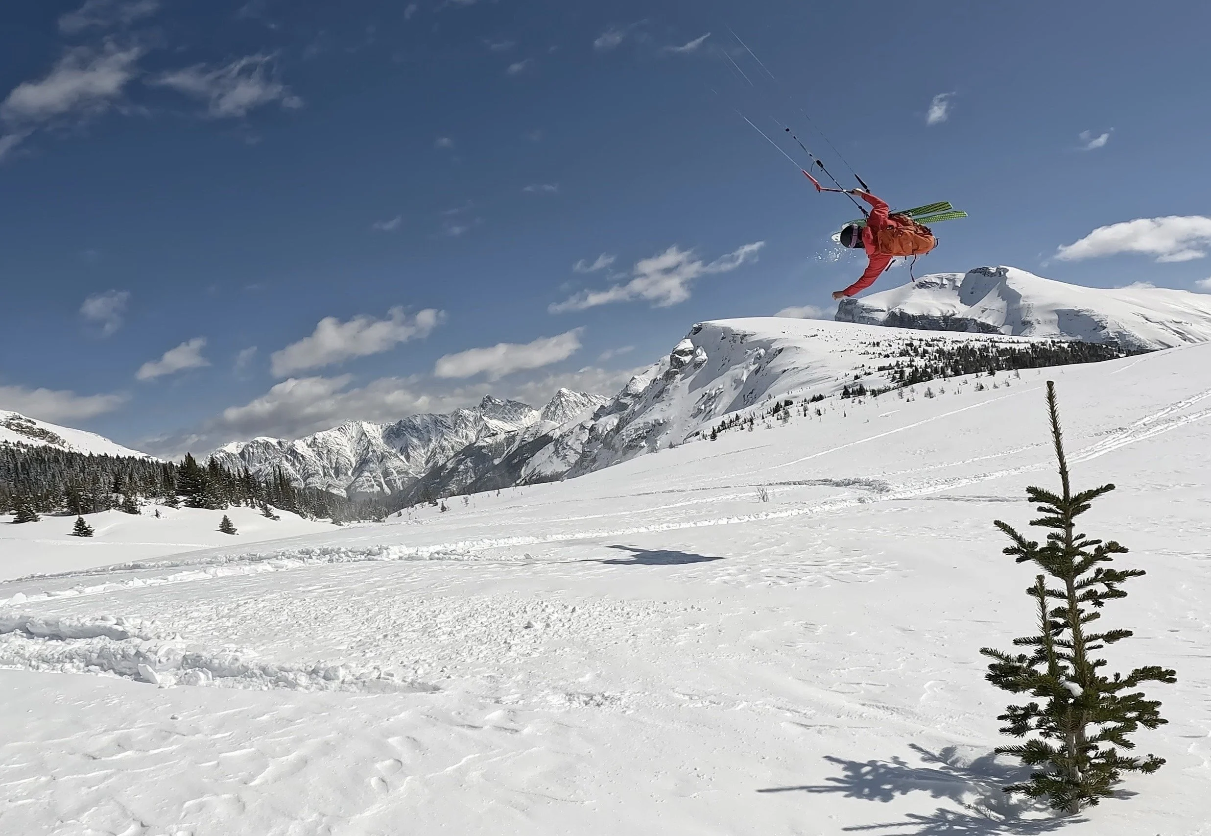 A person falling off a snow-covered mountain while skiing or snowboarding, with mountains and a blue sky in the background.