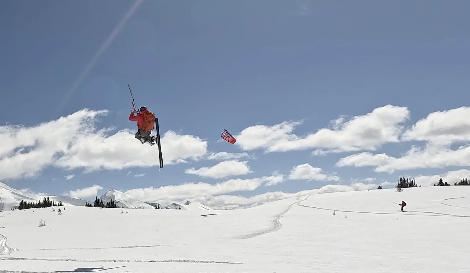 A person in a red jacket and black pants performing a snowboard jump in a snowy mountain landscape with cloudy sky and another snowboarder in the distance. Sunshine Meadows.