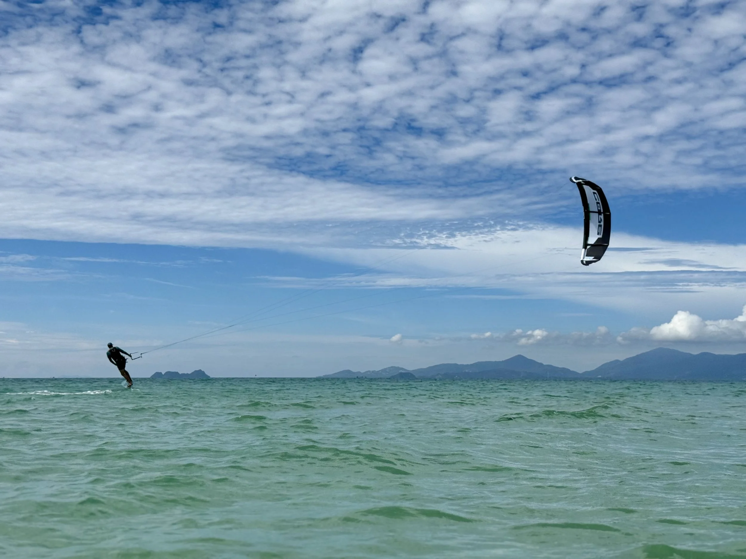 A person kitesurfing on the ocean with a black and white kite, with mountains visible in the background and partly cloudy sky.
