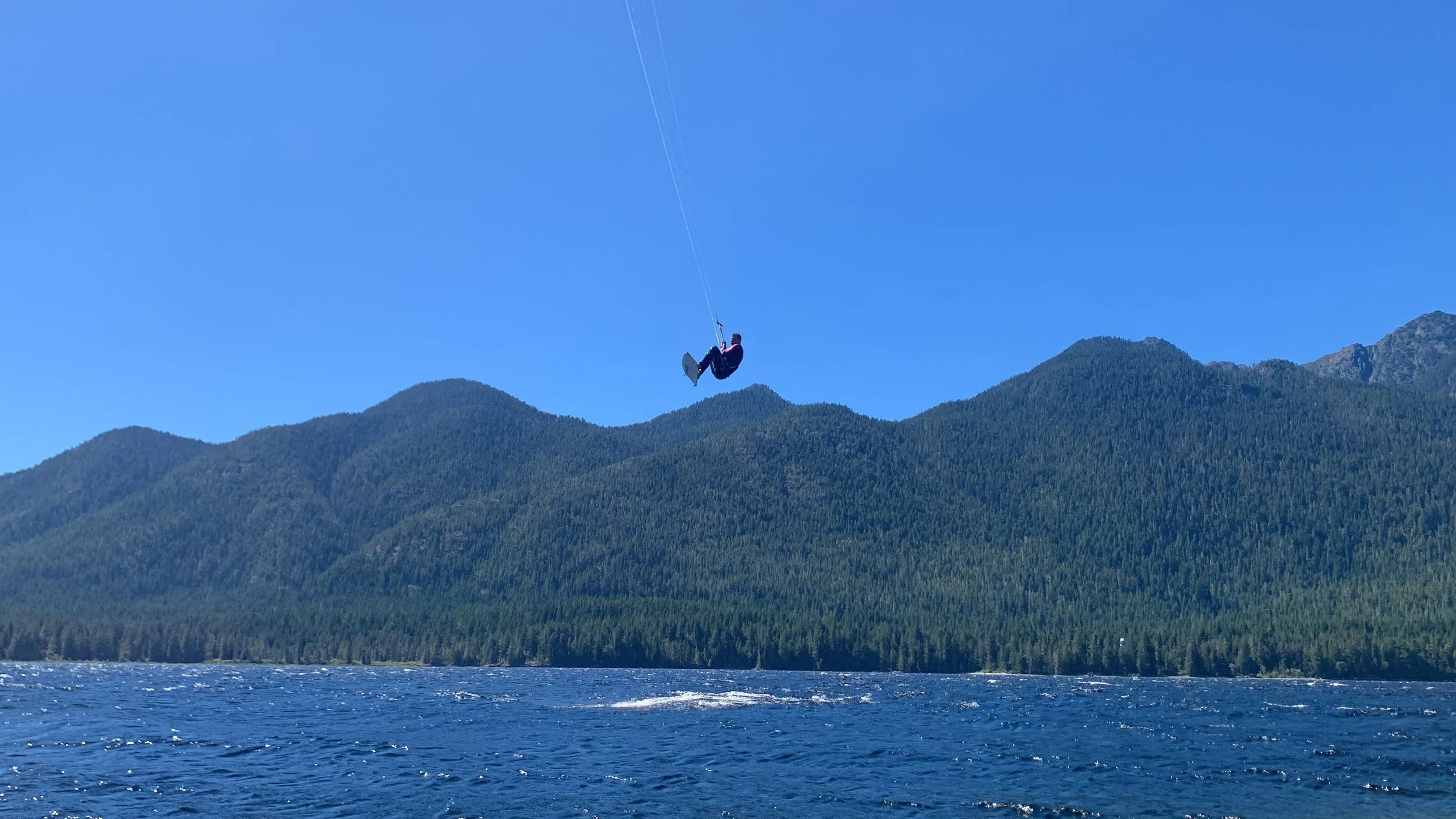 Person flying a foil kite above a body of water with mountains in the background under a clear blue sky.