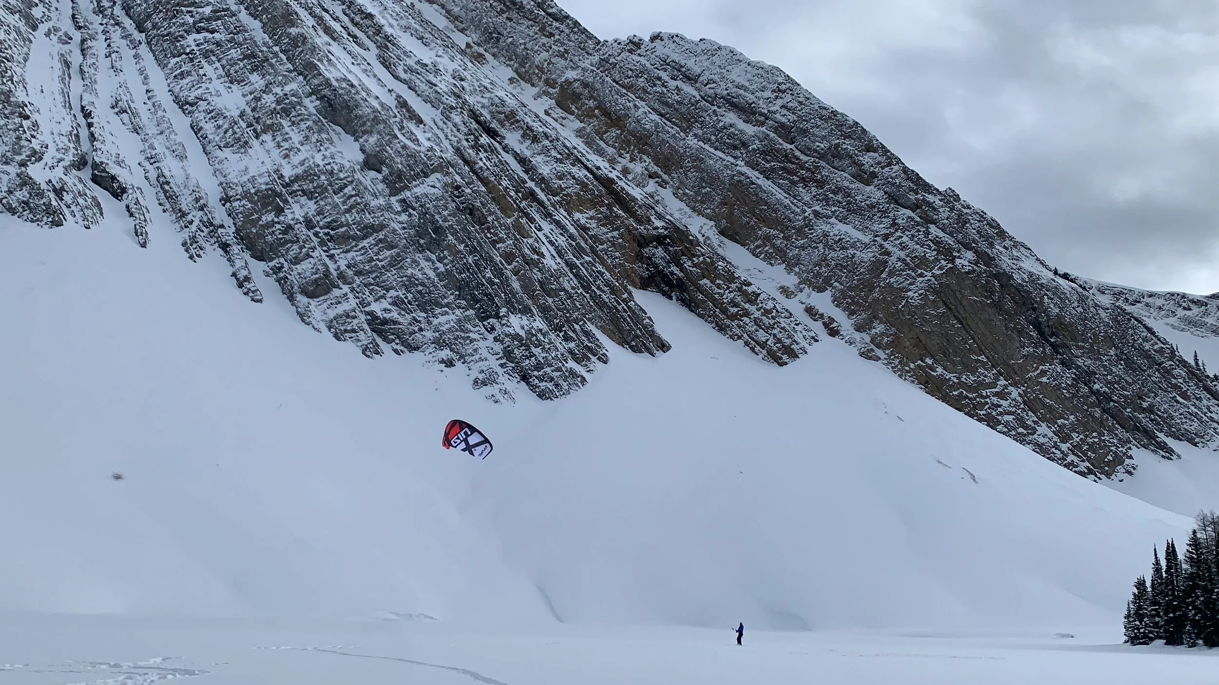 A person in winter clothing stands in a snow-covered landscape, flying a large kite near a steep, snowy mountain with rocky cliffs. Snow kiting Bow Lake, Alberta. Banff National Park. 