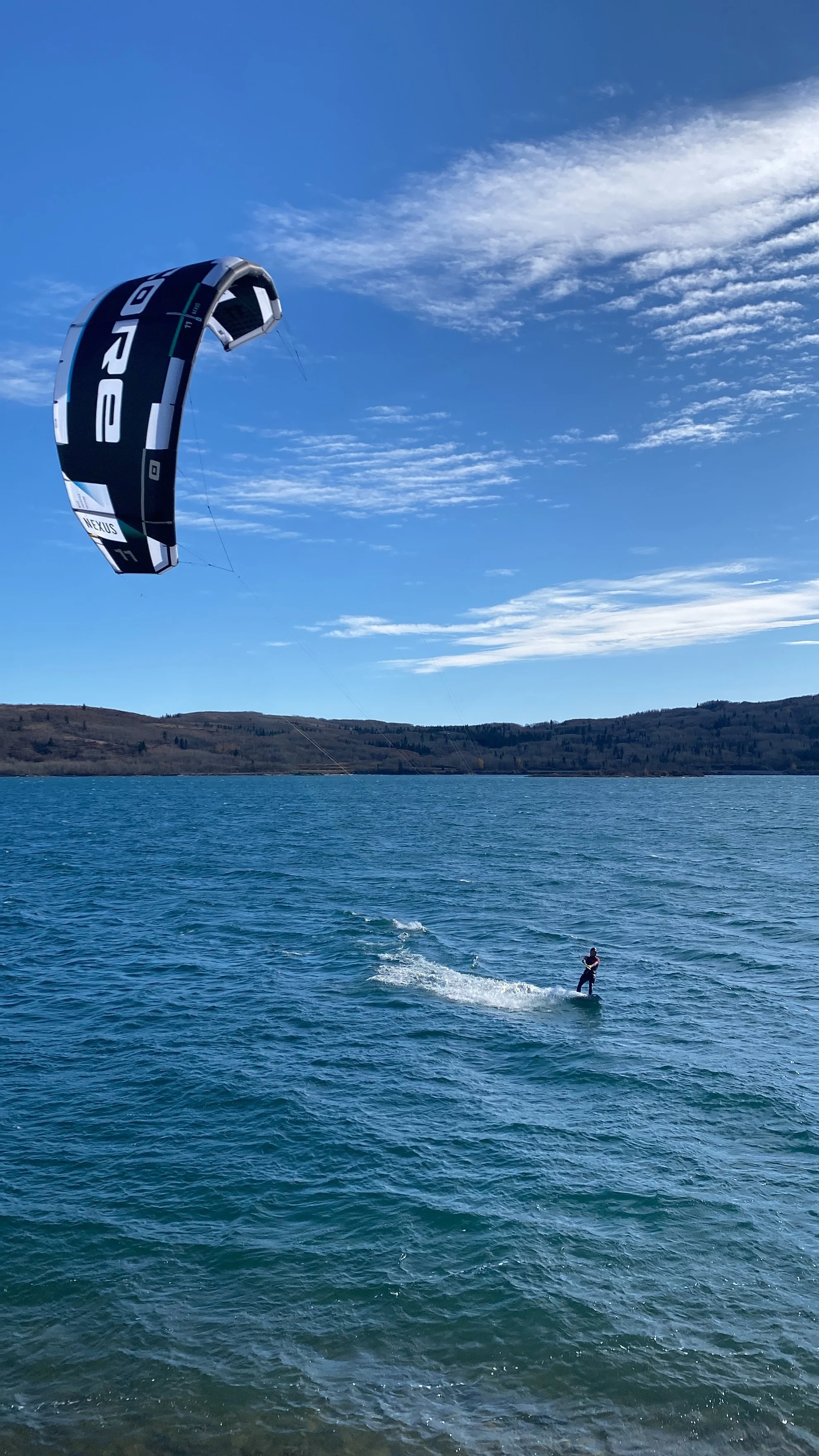 A person kiteboarding on a lake with hilly terrain in the background and a clear blue sky.