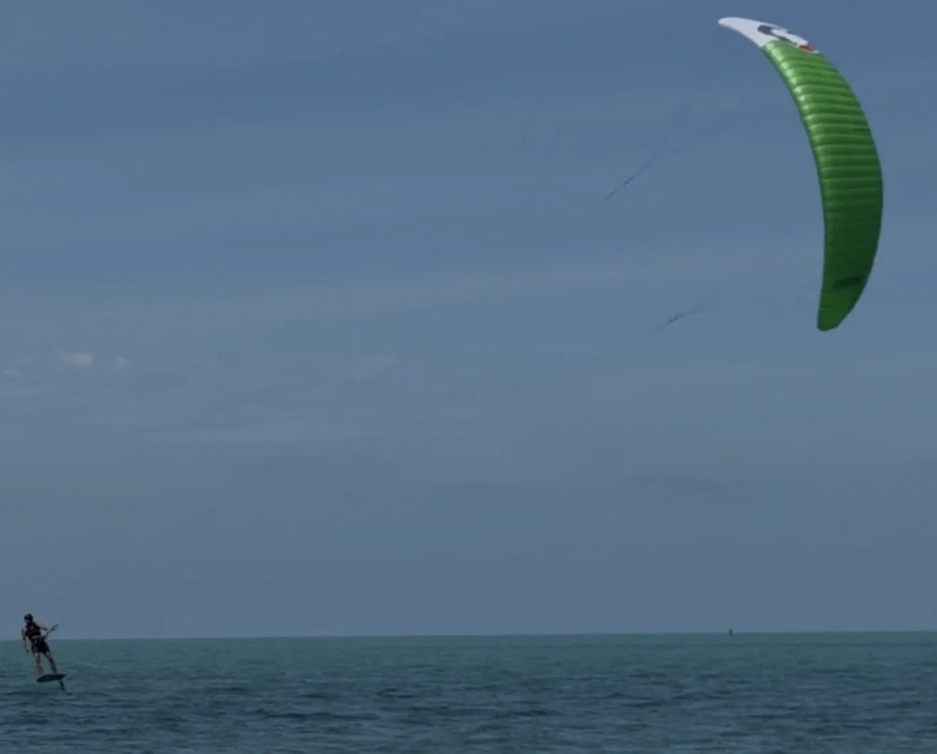 A person on a hydroflight device flying above the water while a kite with a green canopy flies in the sky.