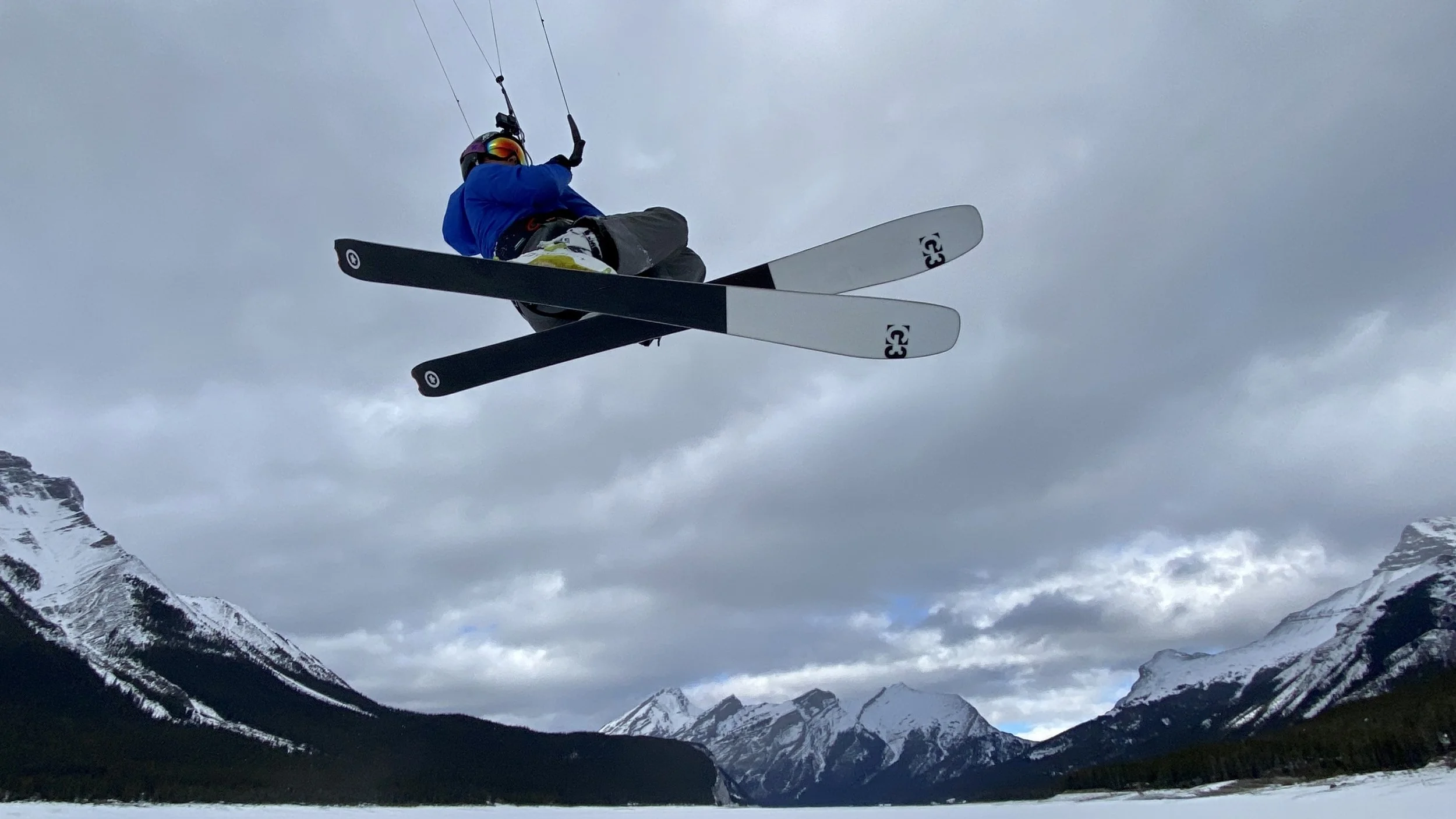 A snowkiter wearing a blue jacket, black helmet with rainbow visor, and skiing gear, is in mid-air performing a jump with snow-covered mountain peaks and a cloudy sky in the background. Riding a Gin Shaman 3.1. Spray Lakes Alberta.
