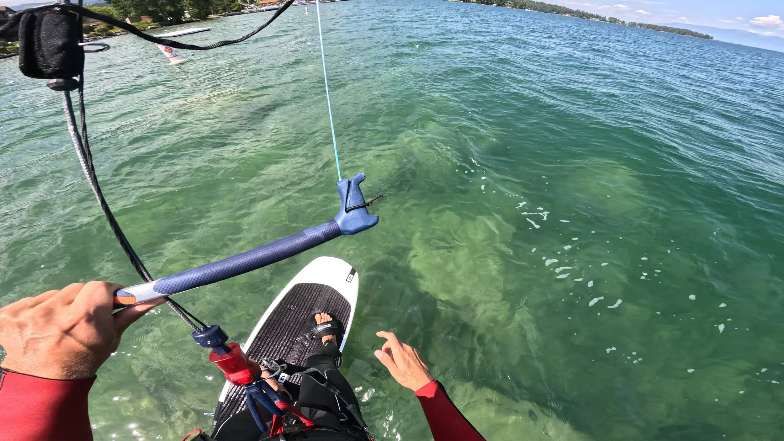 First-person view of a person on a paddleboard in clear green water, holding a fishing rod with a hand, wearing black sandals, with a shoreline and trees visible in the background.
