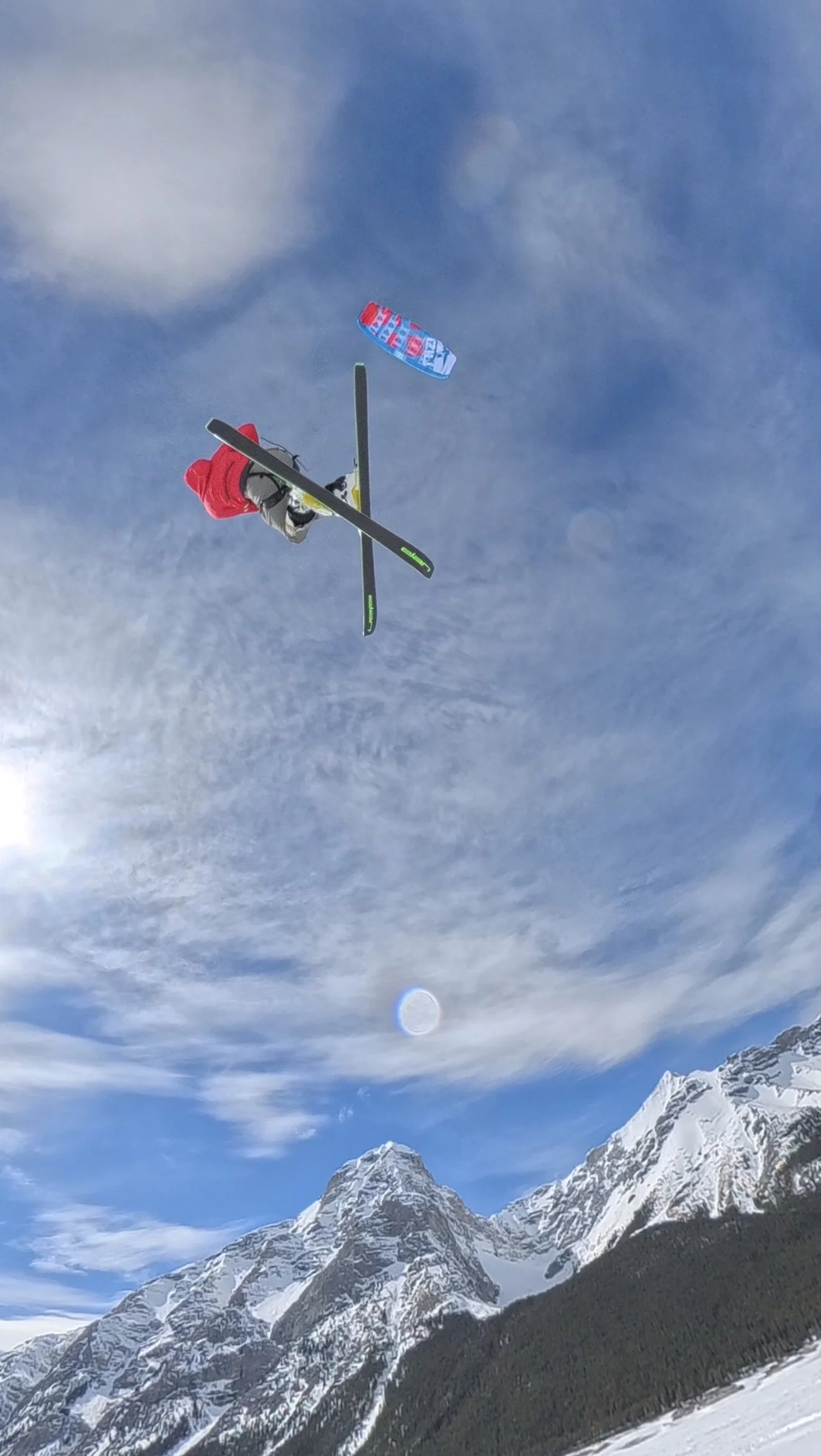 A snow kite skier performing a mid-air stunt against a backdrop of snow-capped mountains and a partly cloudy sky.