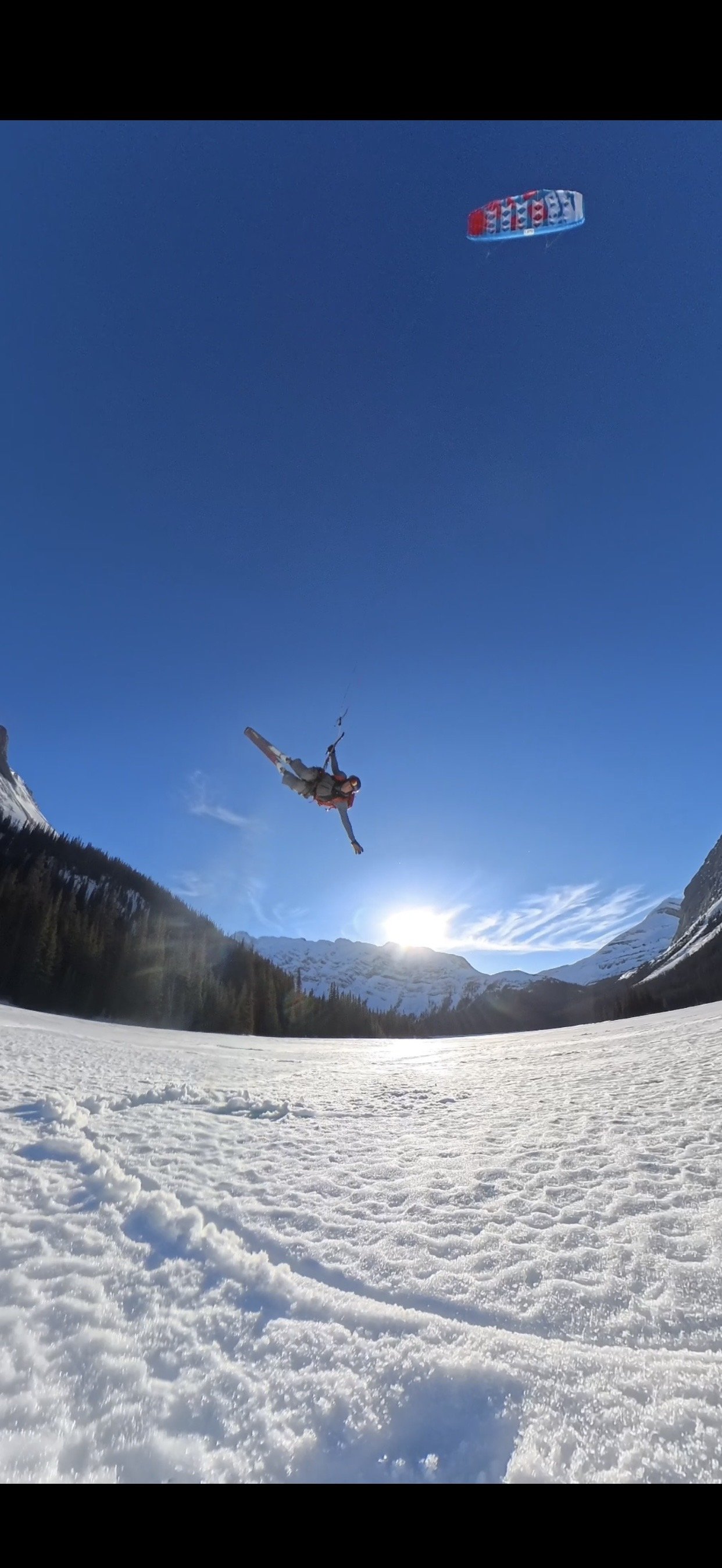 A person snowkiting in a snowy mountain landscape with clear blue sky and the sun low on the horizon.