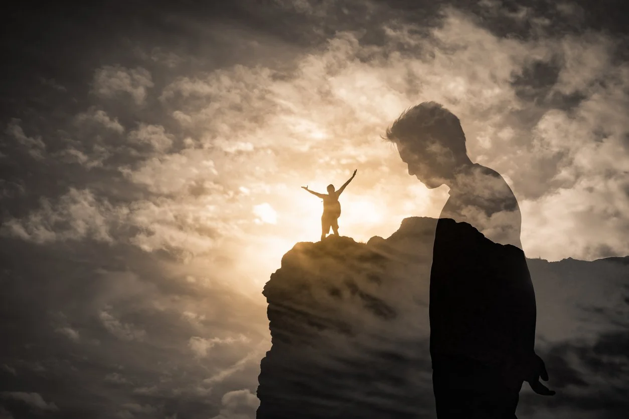 Man standing victorious at the top of a mountain with hands raised to the cloudy sky and in the forefront is the profile of the same man standing with his head slightly bowed.