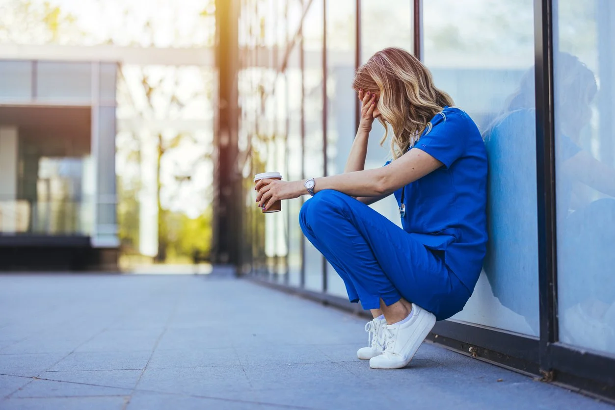 Medical first responder squatting outside with her back to a building, holding a cup of coffee in one hand with her other hand on her face looking down.