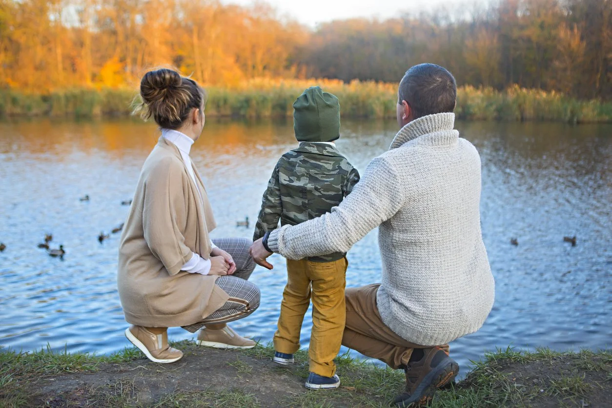 Husband and wife squatting beside a lake with their son in the middle looking at the ducks.
