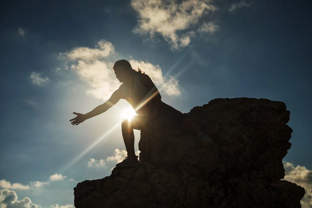 Woman at the top of a mountain with the sun shining in the back as she reaches her hand down to help pull someone up from grief and loss.