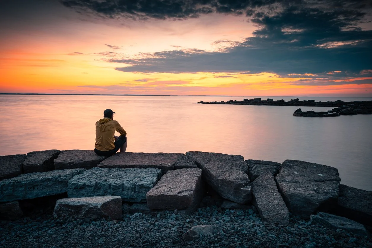 Man sitting on rocks near downtown Burlington watching the sun set over Lake Ontario.