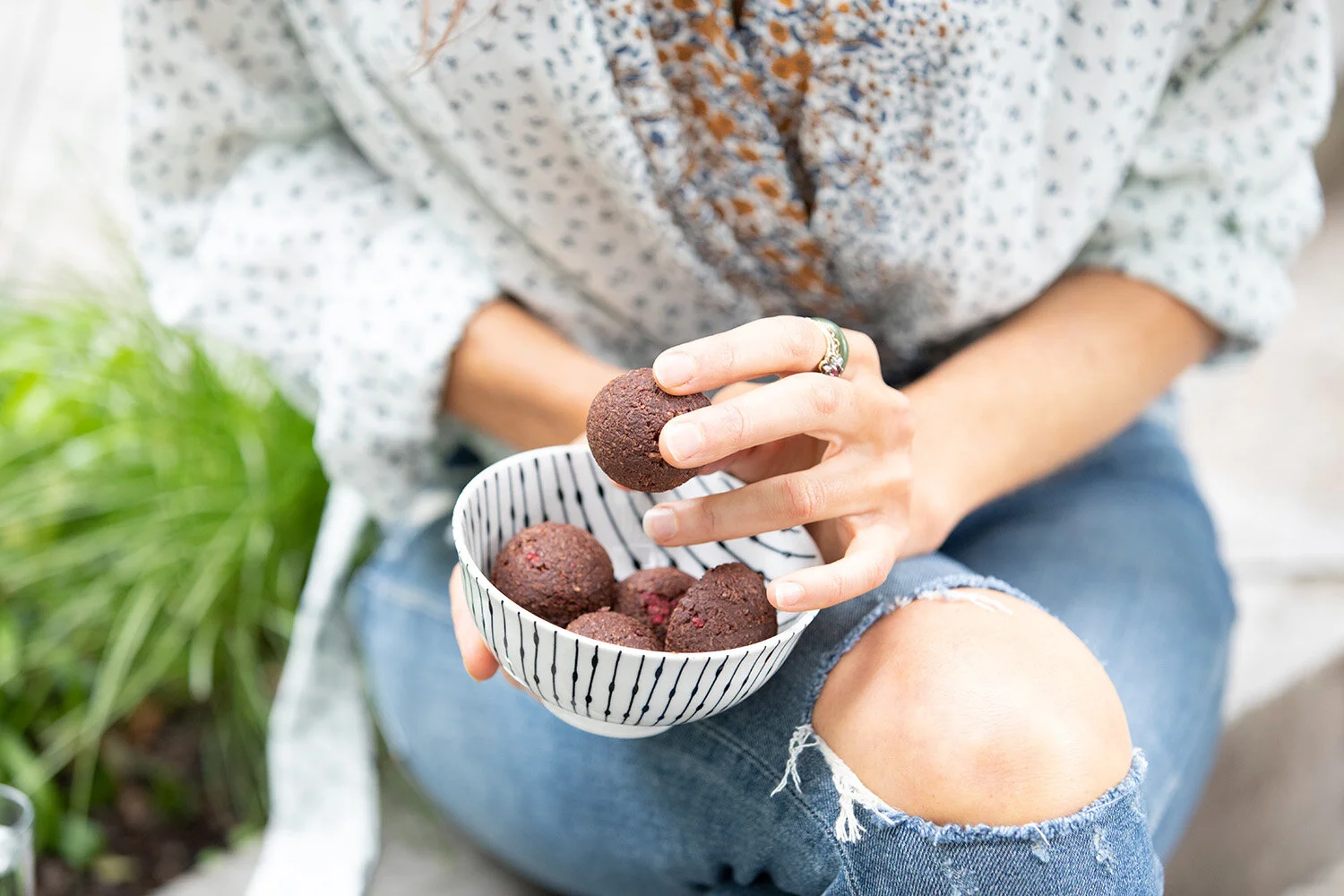 Person holding a striped bowl filled with chocolate truffles, reaching for a truffle, seated outdoors.