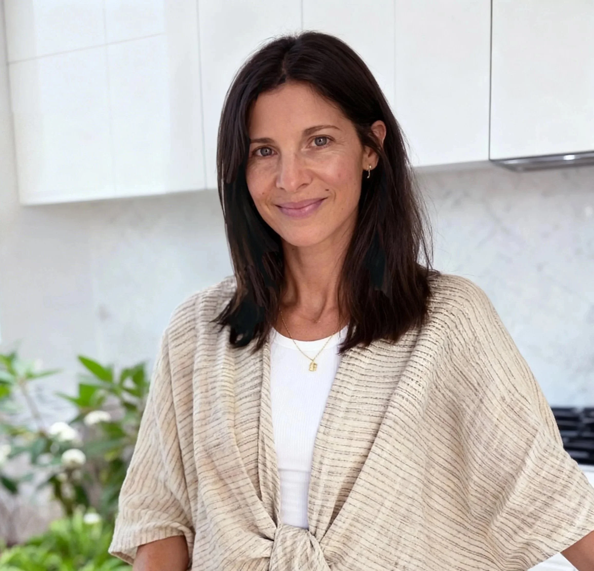 A woman with shoulder-length dark brown hair, smiling, wearing a beige tie-front cardigan over a white top, with a small gold necklace, and standing in a bright kitchen with white cabinets and green plants in the background.