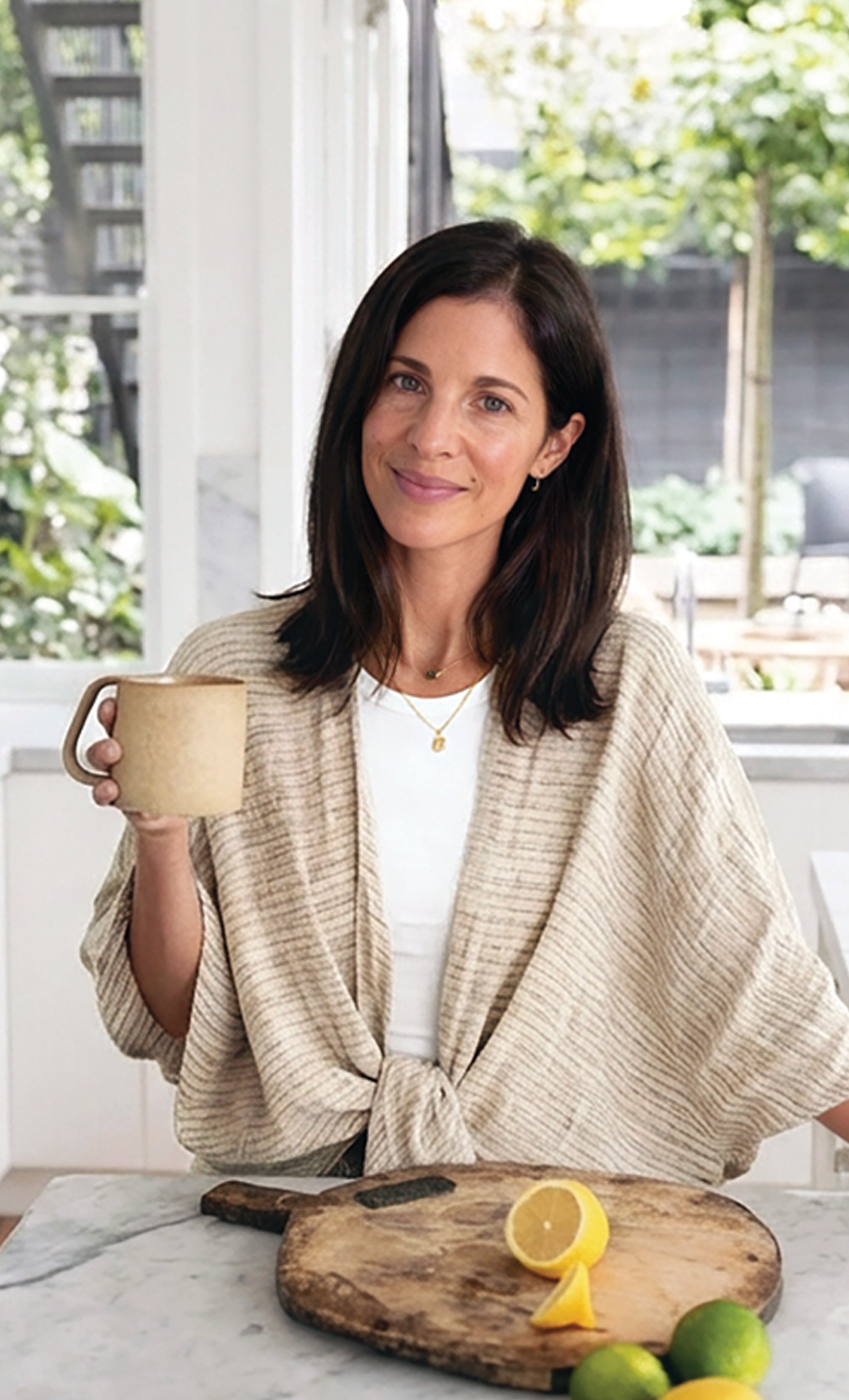 A woman with dark brown hair and light skin, wearing a beige shawl and white shirt, holding a mug, standing in a bright kitchen with a lemon on a cutting board and limes on the counter.