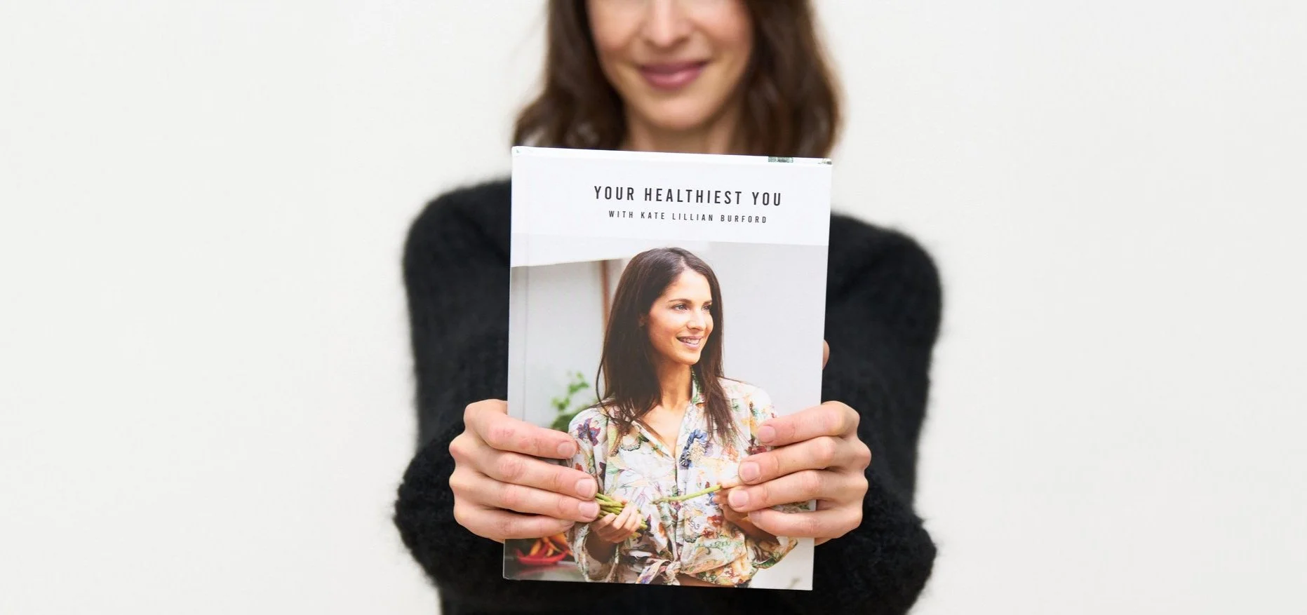 A woman with brown hair and a black sweater holding a magazine titled 'Your Healthiest You' featuring a smiling woman with dark hair wearing a floral blouse on the cover.
