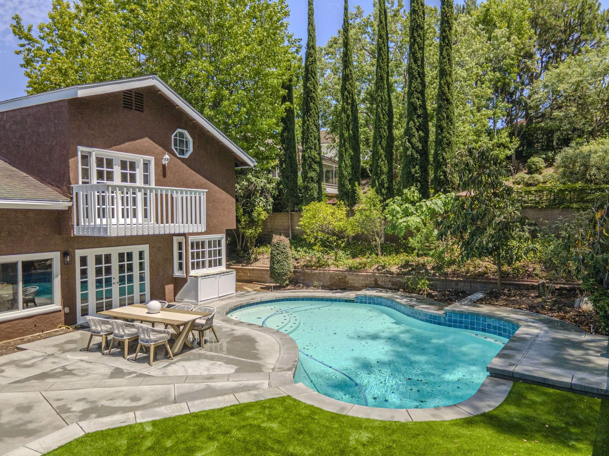 Living room with fireplace, wooden mantel, flat-screen TV, large windows showing a green yard, gray sofa, black coffee table with a vase of flowers, potted plant, mirror on wall, wooden side table, decorative pillows, and a rug.