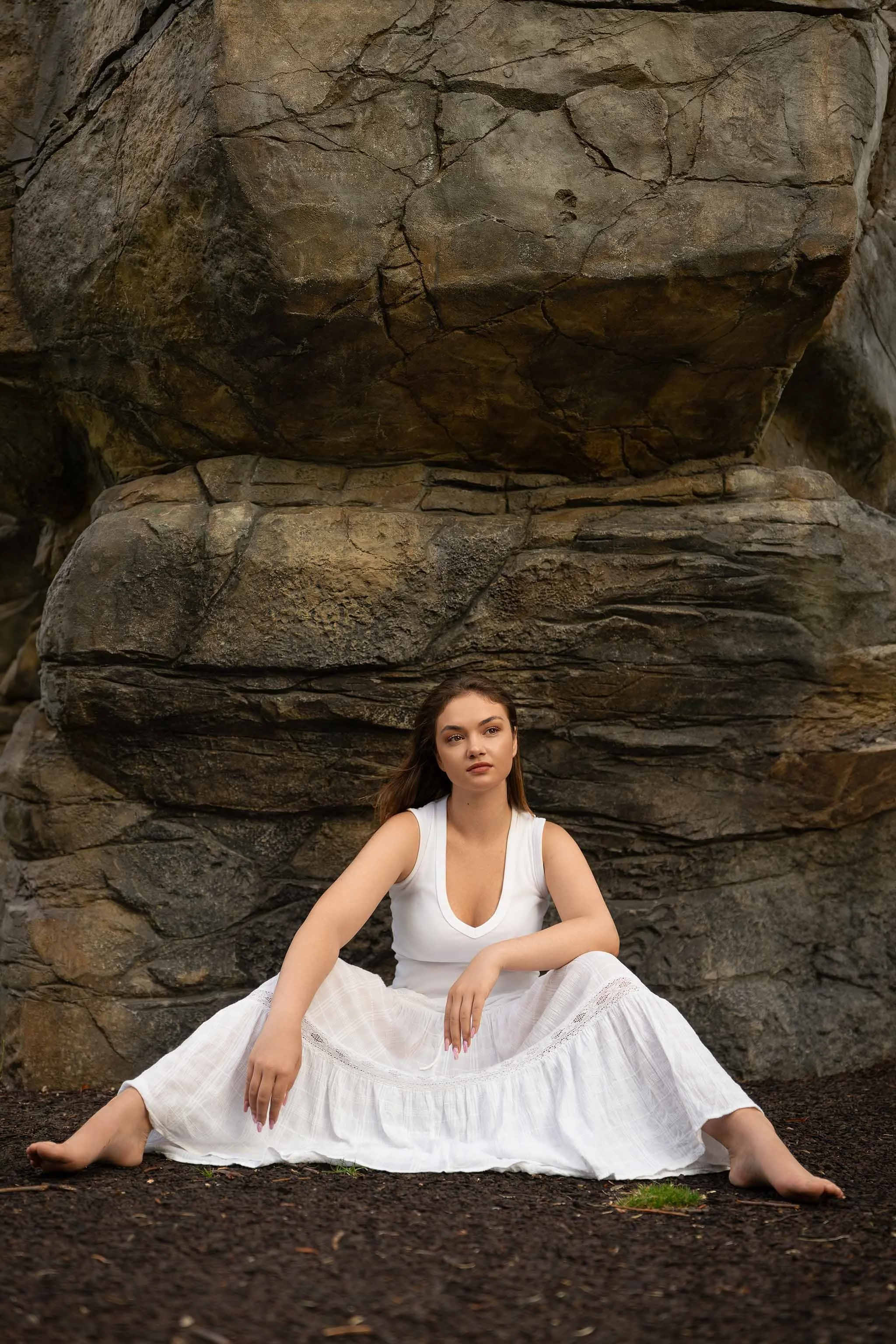 Grounded and serene outdoor portrait of a barefoot woman in white, seated in front of raw stone textures — evoking strength, stillness, and natural beauty.