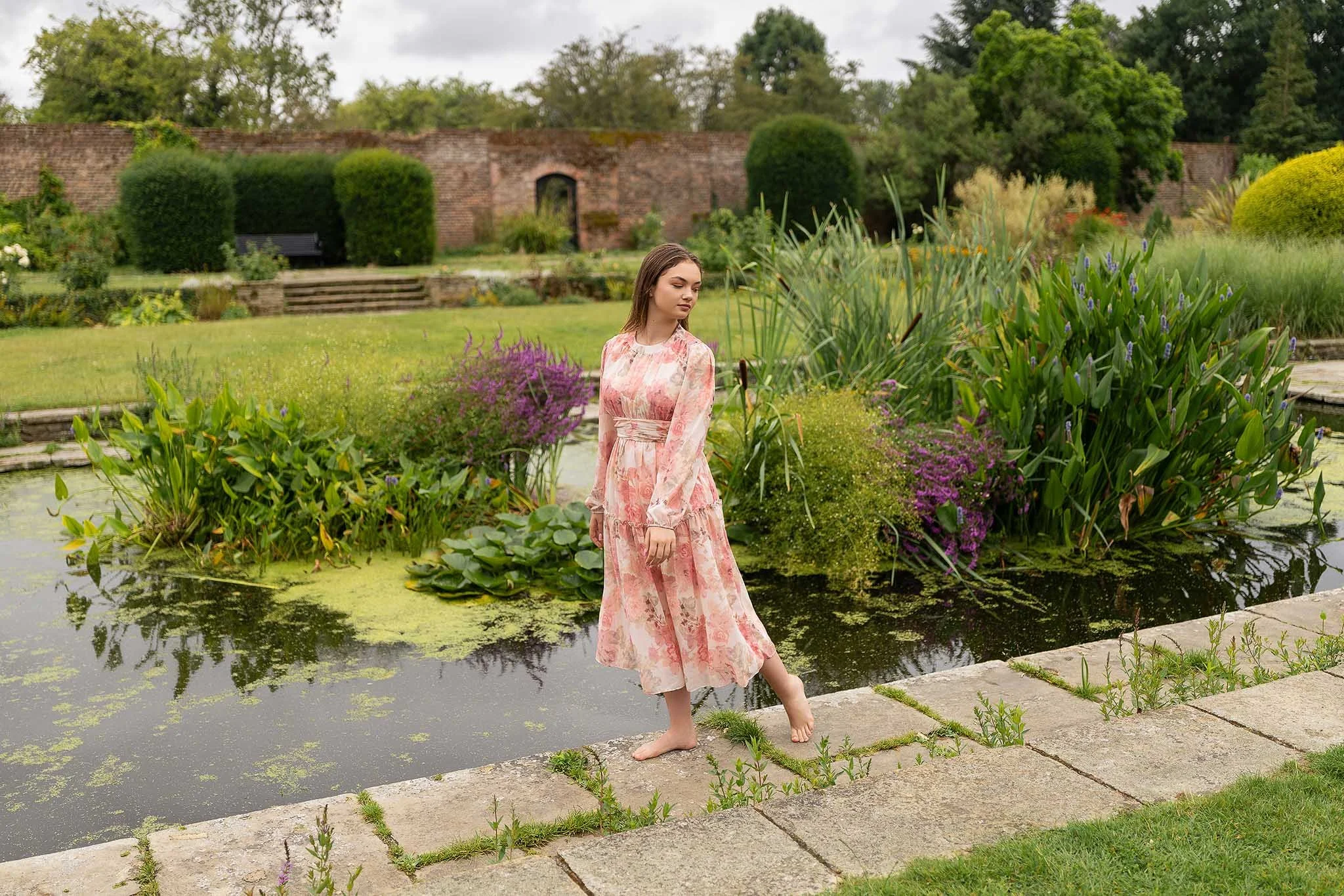 A woman in a pink floral dress is walking through a stone garden path. Calm, graceful atmosphere captured in natural light.