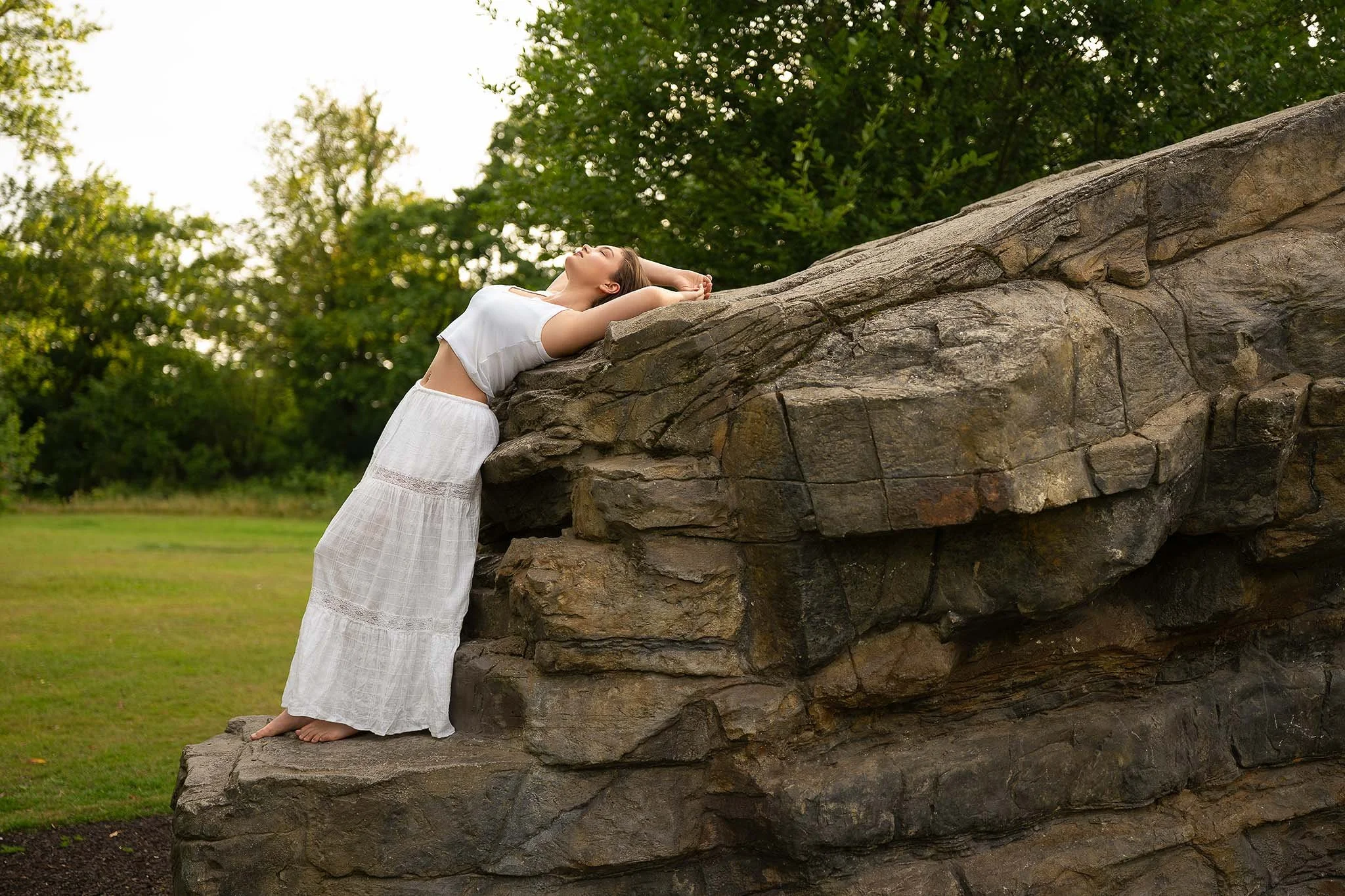 Ethereal outdoor portrait of a barefoot woman in a white flowy outfit, arching gracefully on a rock ledge — radiating freedom, femininity, and harmony with nature.