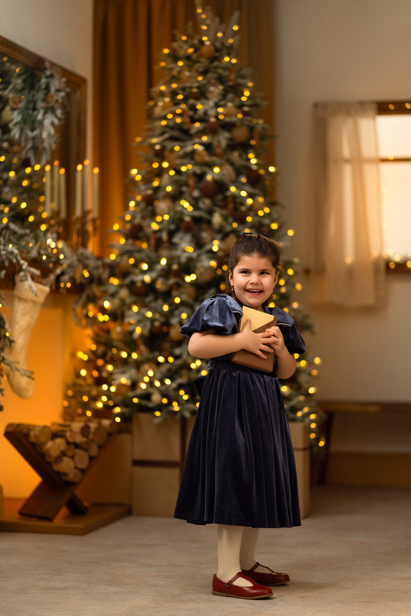 Little girl in a navy velvet dress holding a gift beside a glowing Christmas tree in a warm indoor Christmas set.