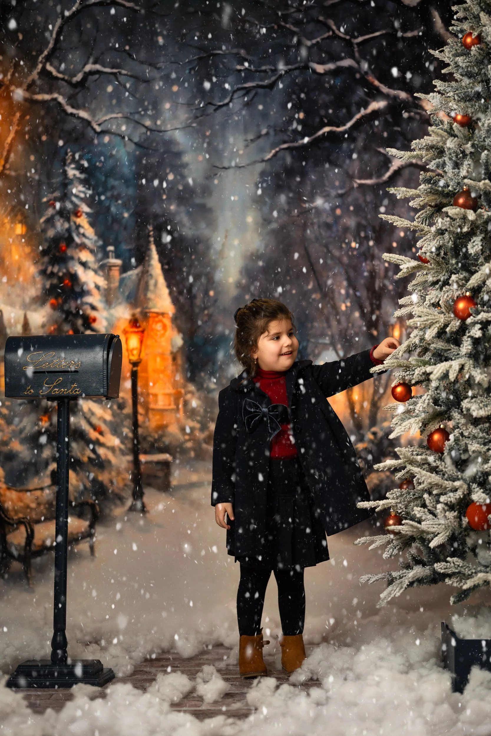 Little girl reaching for a red bauble beside a snow-covered Christmas tree, with falling snow and a Letters to Santa postbox in the winter outdoor studio set.