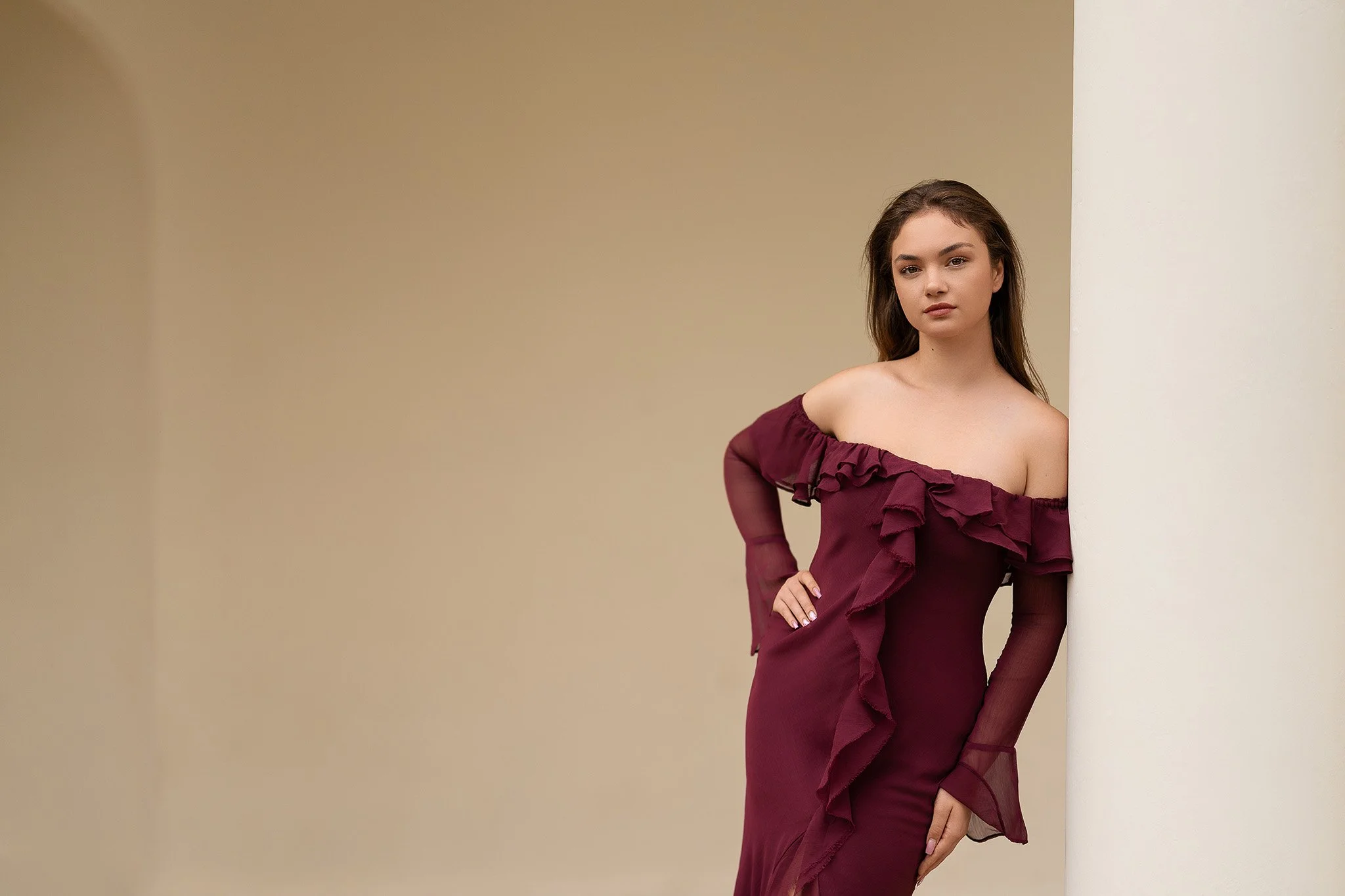 Refined outdoor portrait of a woman in a burgundy off-shoulder gown, captured against a smooth neutral wall and classic white column for an elegant, timeless look.